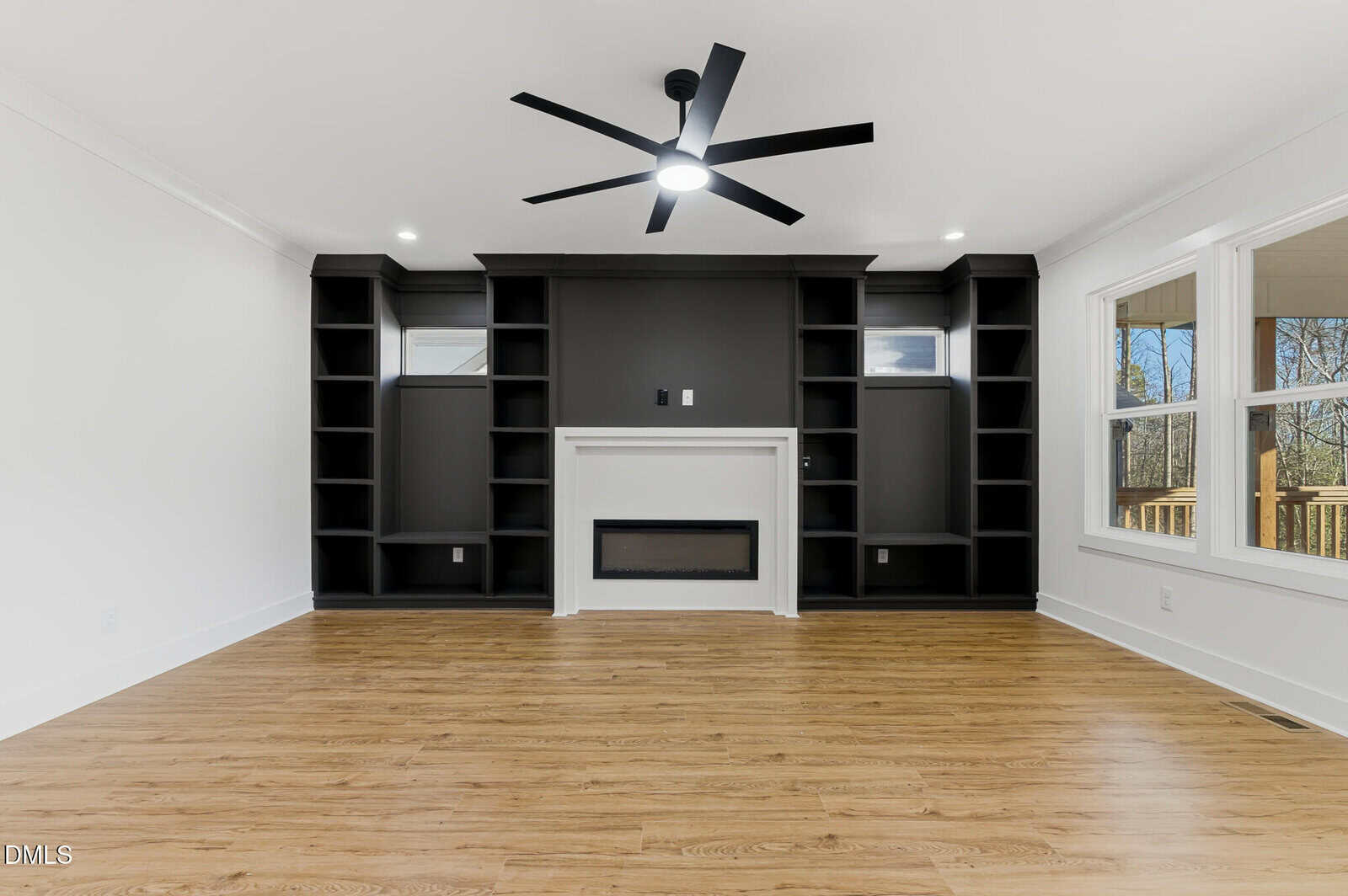 Modern living room with light wood floors, a black ceiling fan, and a black accent wall featuring built-in shelves and a white fireplace. Large windows on the right let in natural light.
