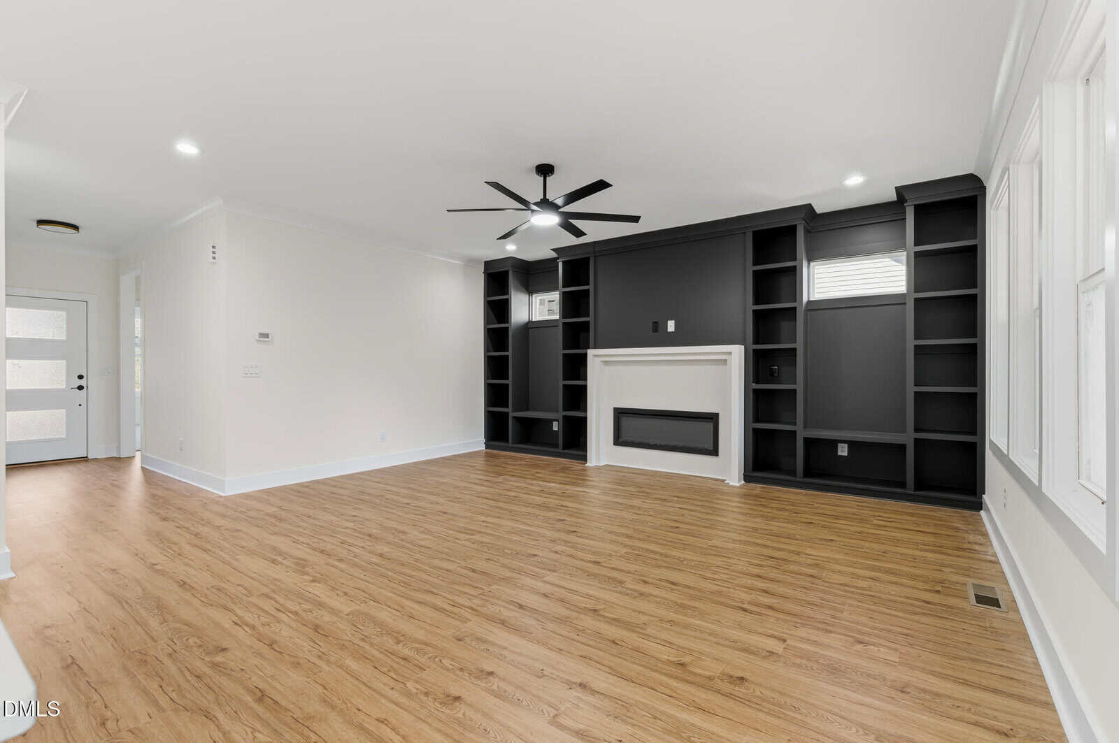 A spacious, modern living room with light wood flooring, white walls, a black ceiling fan, and built-in black shelving around a white fireplace. Large windows on the right wall let in natural light.