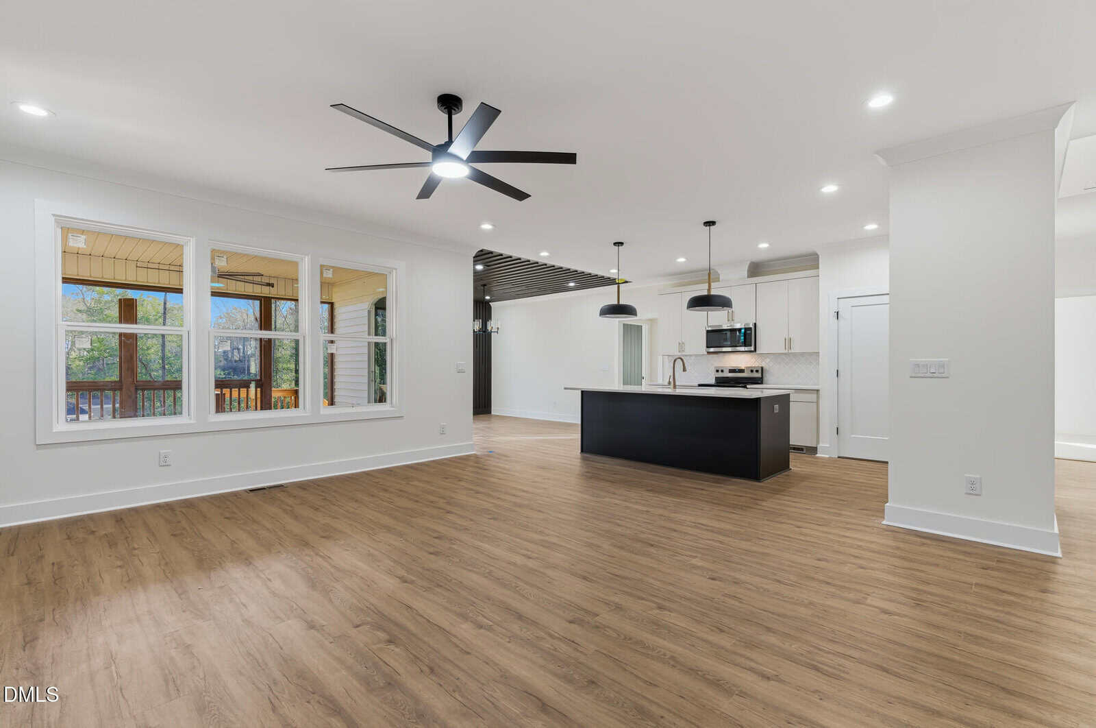 Modern open-concept living area with wood flooring, large windows, a black ceiling fan, and a kitchen featuring a black island, pendant lights, and stainless steel appliances. Covered porch visible through the windows.