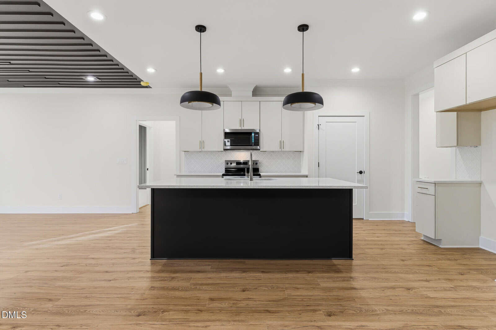 Modern kitchen with wood floors, a large black island, white cabinets, stainless steel appliances, two black pendant lights, and recessed ceiling lights in a bright, open space.