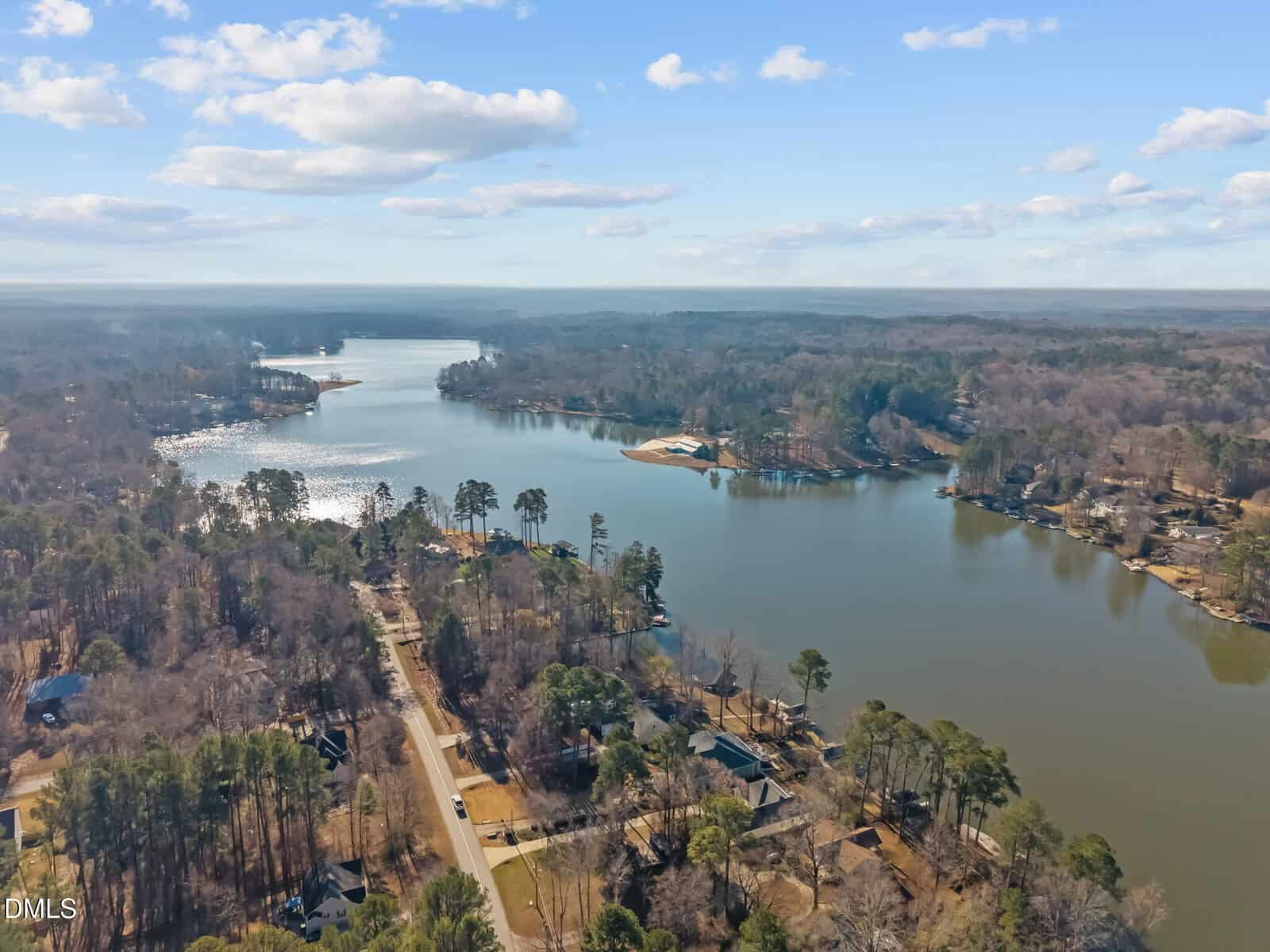 Aerial view of a peaceful lake surrounded by trees and houses, with roads running alongside the shoreline under a blue sky with scattered clouds.