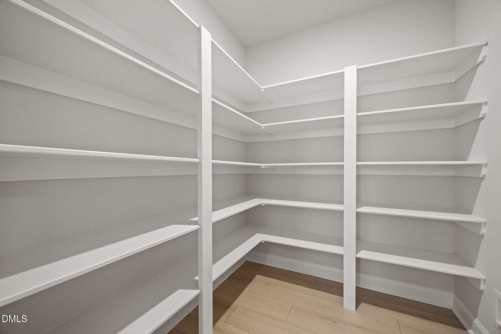 A spacious walk-in pantry with white shelves lining the walls and light wood flooring, all brightly lit and empty.