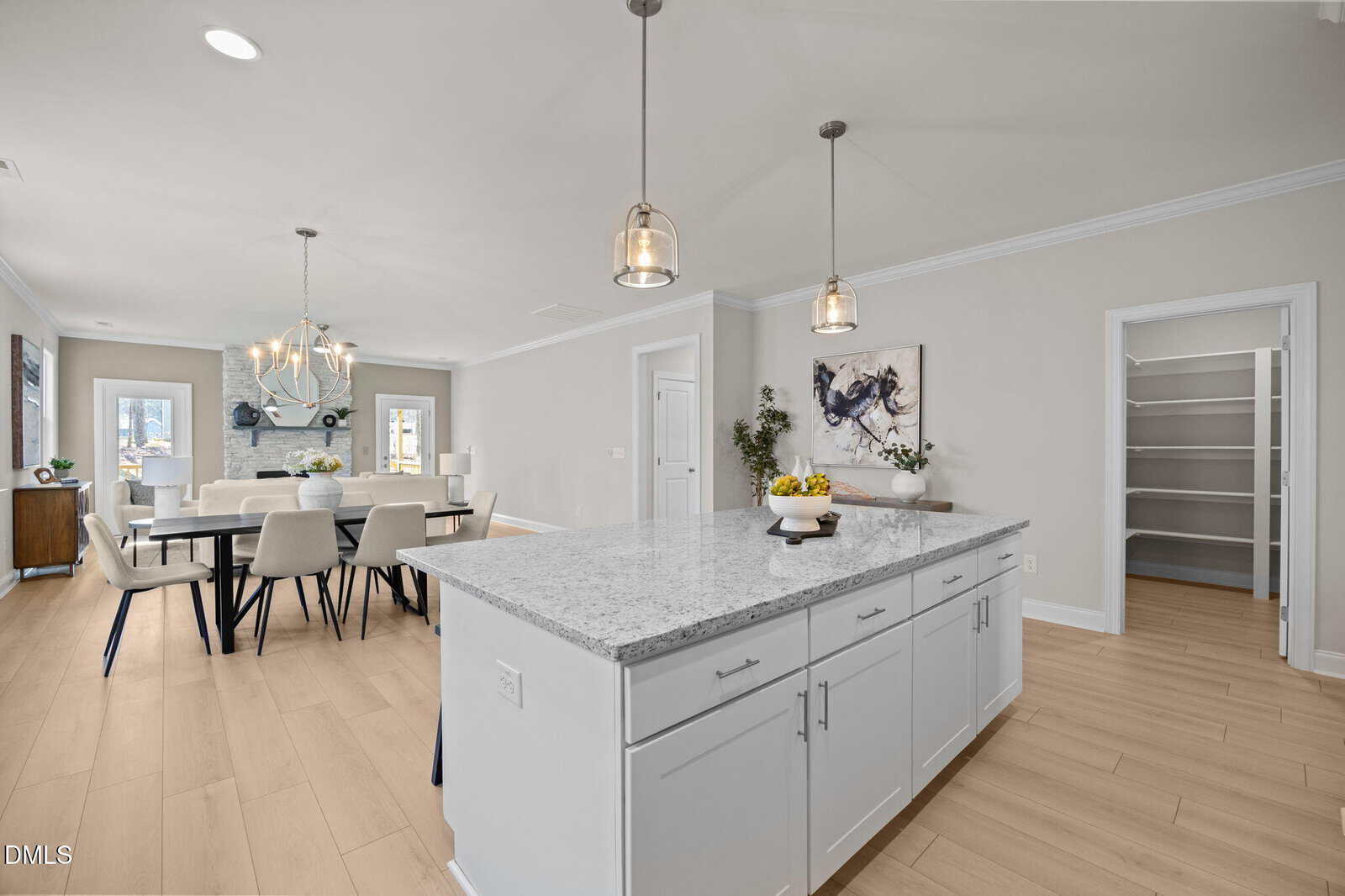 Modern open-concept kitchen and dining area with light wood floors, a large white island with pendant lights, and a spacious pantry. Dining table with white chairs sits near a cozy fireplace and large windows.