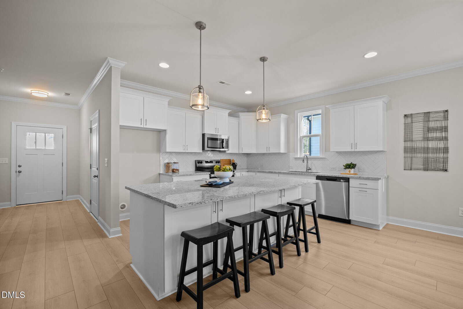 Modern kitchen with white cabinets, stainless steel appliances, marble countertops, a large island with four black stools, pendant lighting, and light wood flooring. A window and wall art add natural light and style.