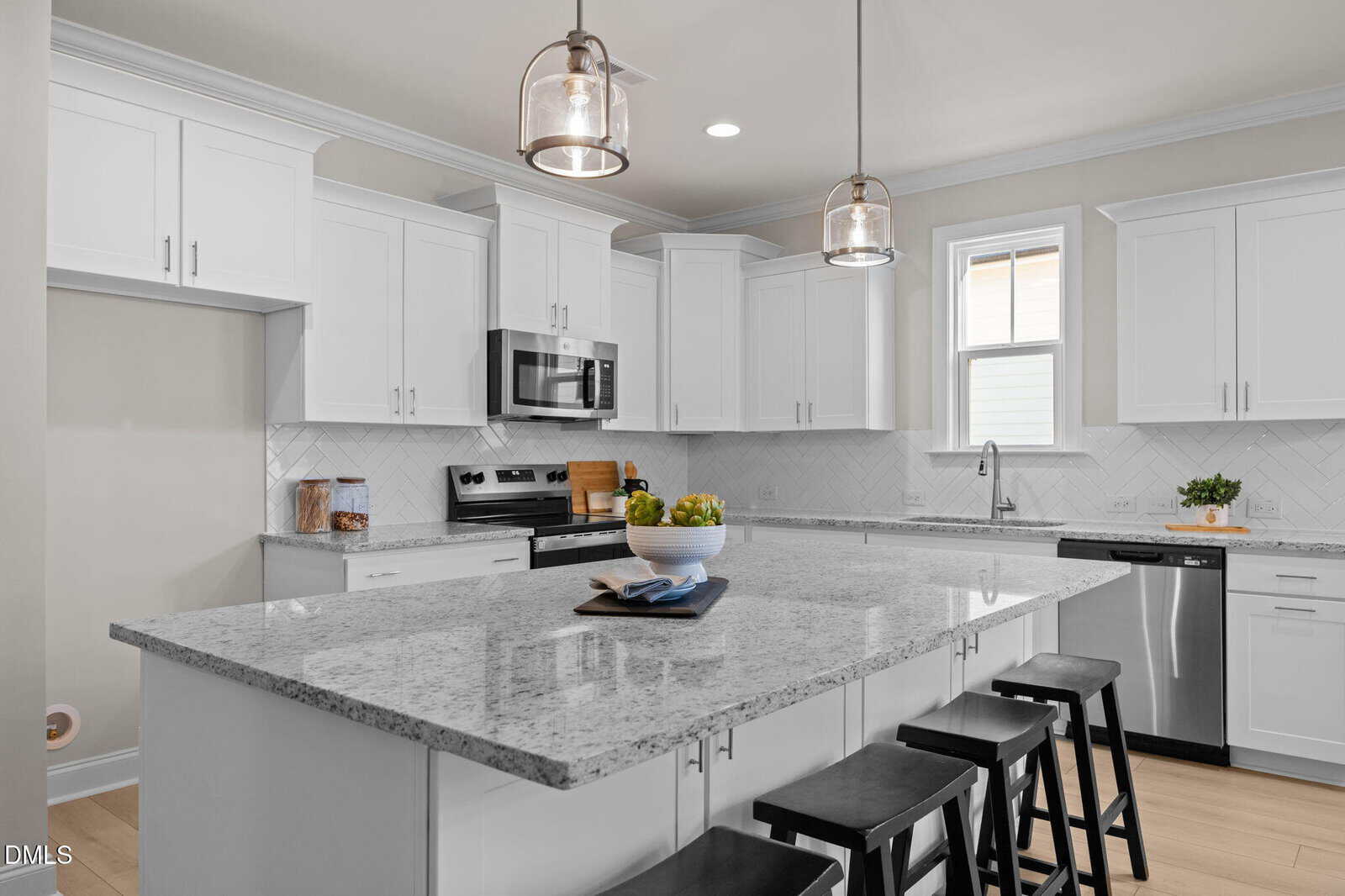 Modern kitchen with white cabinets, stainless steel appliances, a large granite island with black stools, pendant lighting, and a window above the sink. The counter features a bowl of green apples and two stacked books.