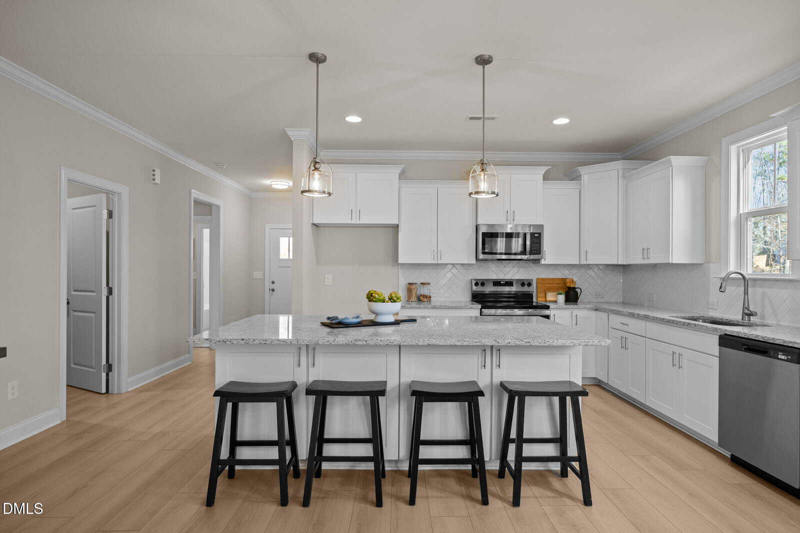 Modern kitchen with white cabinets, stainless steel appliances, marble island with four black stools, pendant lights, wood flooring, and natural light from a window above the sink.