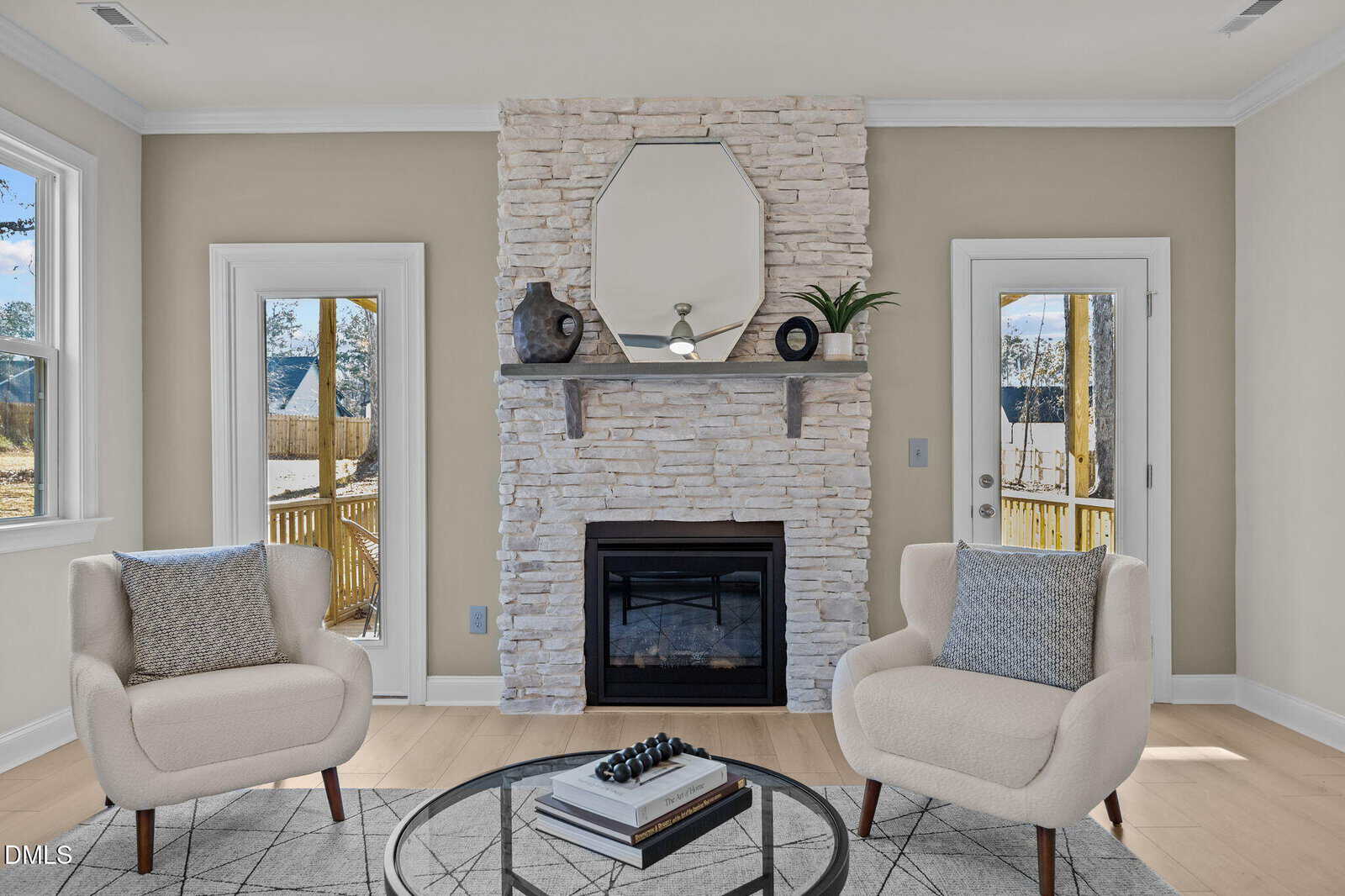 A bright living room with two armchairs, a round coffee table, and a white stone fireplace with a mantel decorated with artwork and vases. Large windows and glass doors let in natural light.