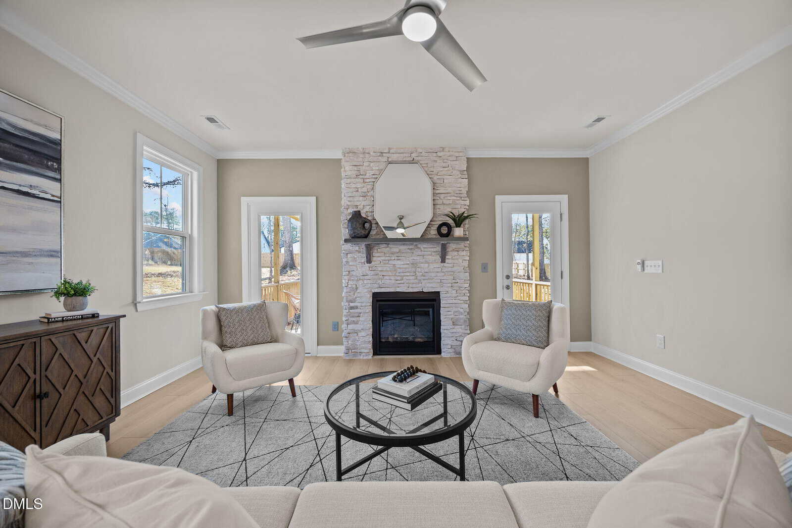 Bright living room with two cream armchairs, a round glass coffee table, and a stone fireplace with a round mirror and decor, flanked by two windows and two glass doors.