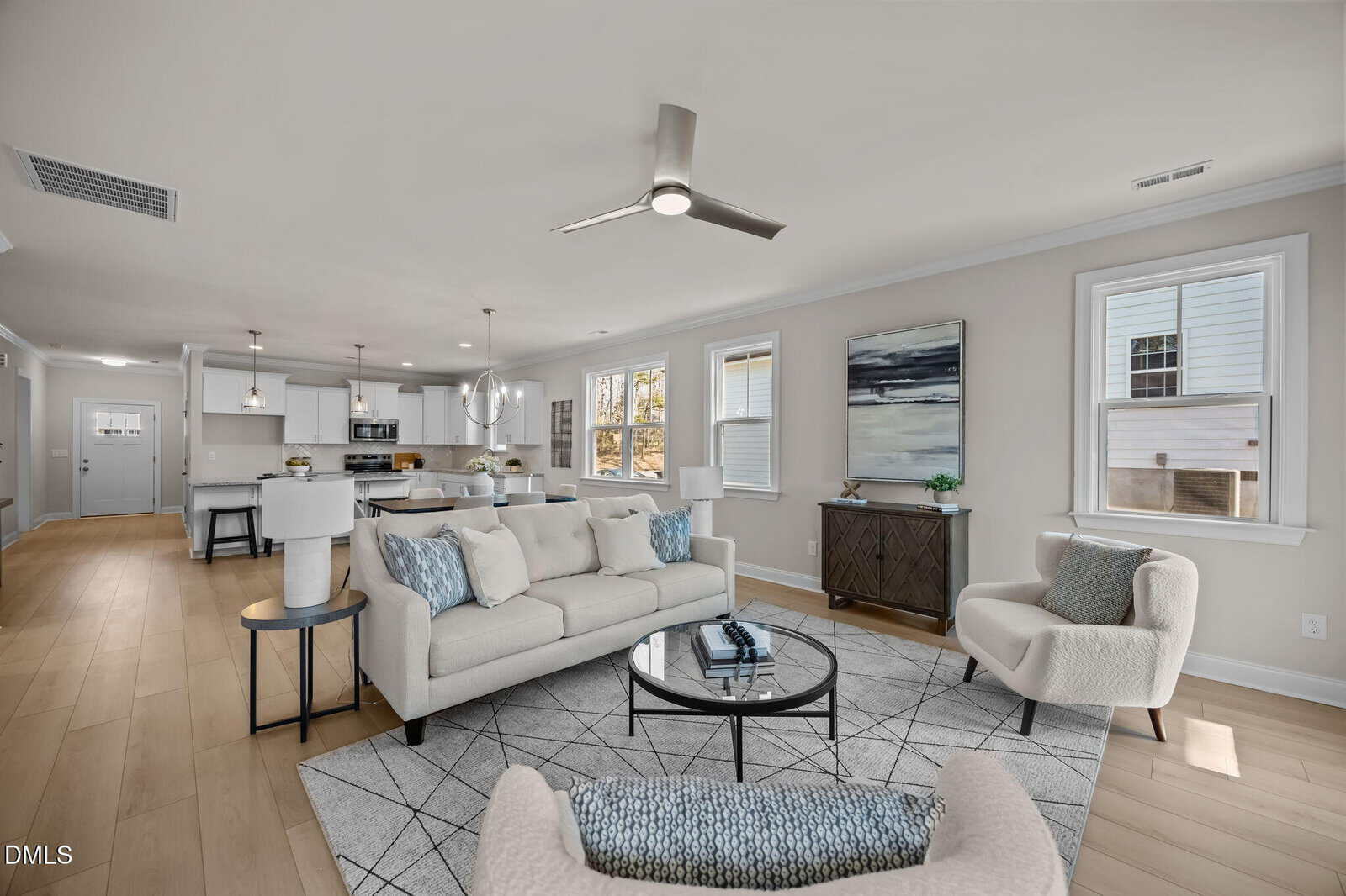 Modern open-concept living room with neutral decor, featuring a white sofa, armchairs, round coffee table, light wood flooring, and a view into a bright kitchen with white cabinets and a dining area. Large windows provide natural light.