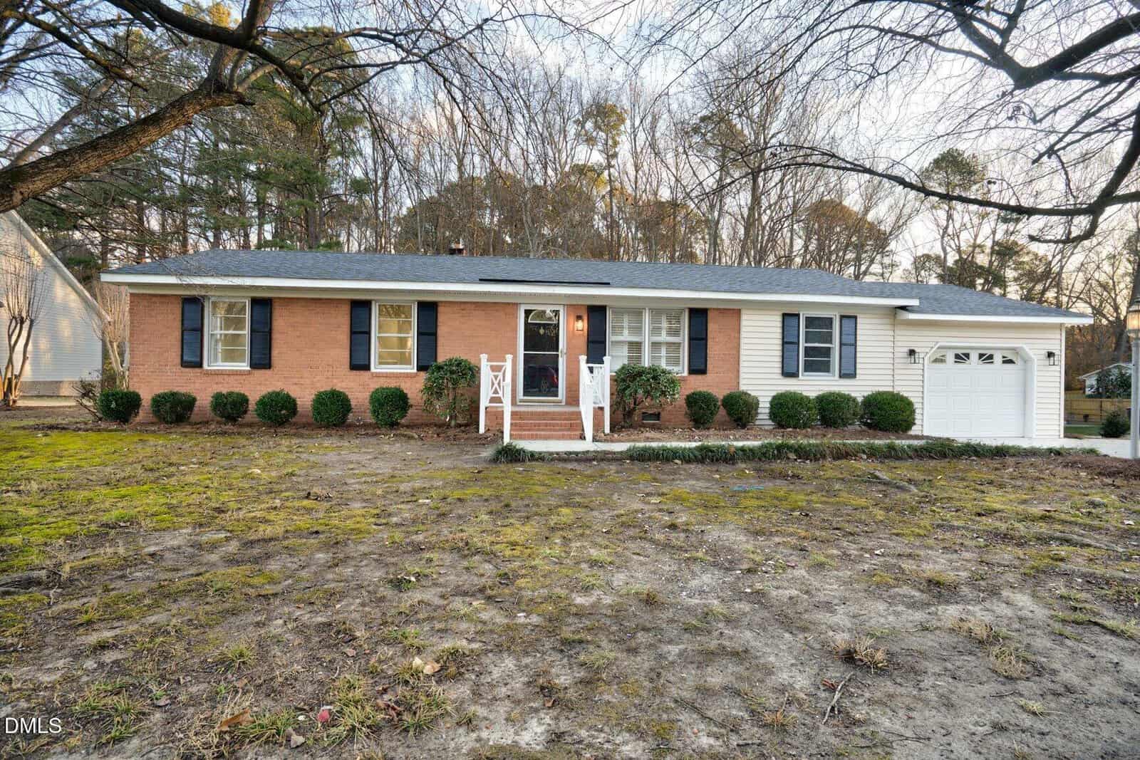 Charming single-story brick house at 201 Drew Street, Louisburg, with dark shutters, white trim, a cozy front porch with railings, and an attached one-car garage. Sparse grass and bushes line the yard, with trees visible in the background.