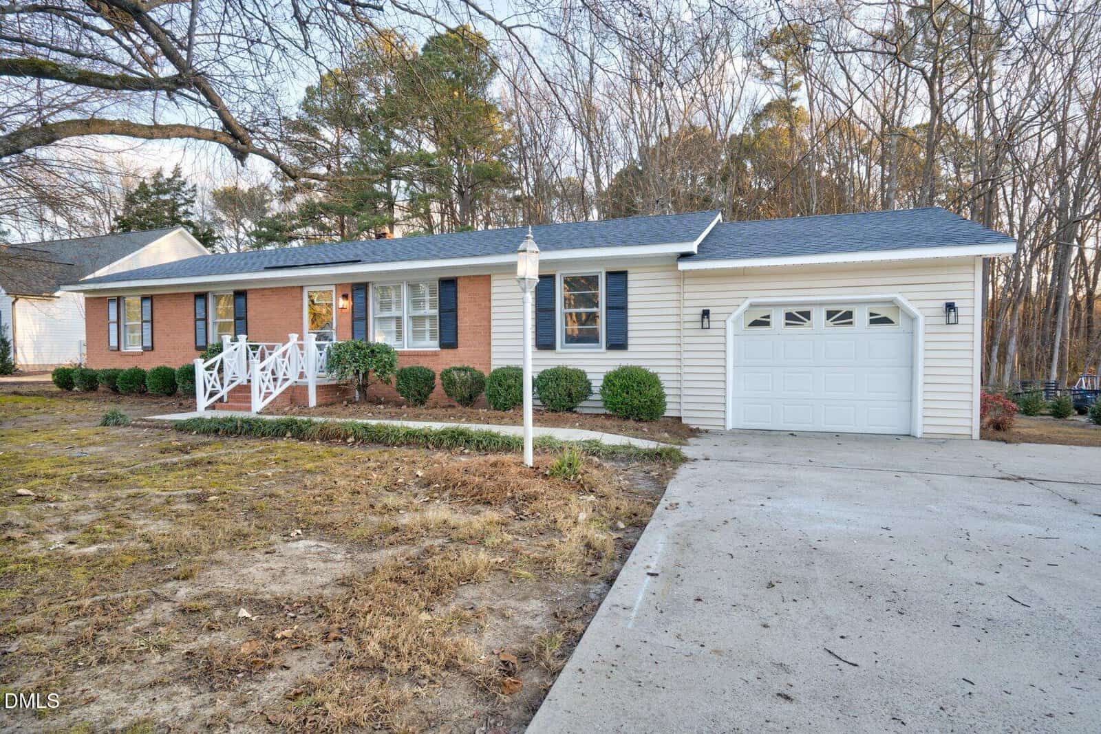 Single-story house with a mix of red brick and light siding, white trim, and a front porch with white railings. There is a one-car garage, a concrete driveway, and neatly trimmed shrubs along the front.