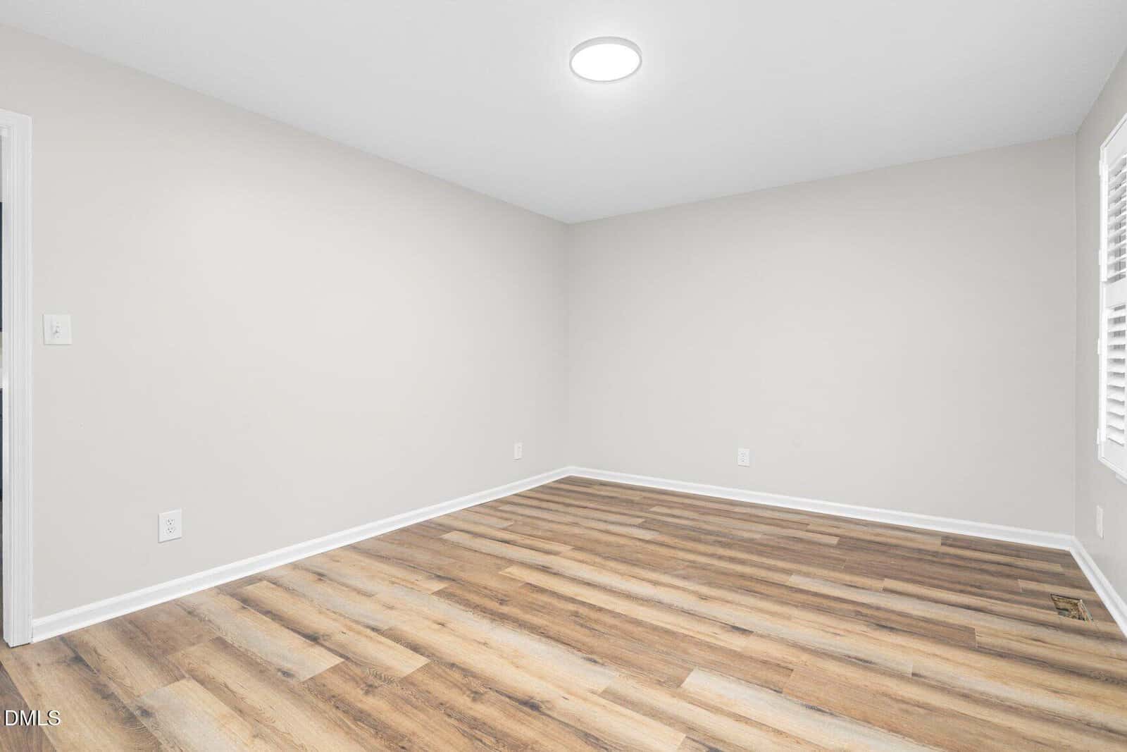 An empty room with light beige walls, wood-style laminate flooring, white baseboards, a round ceiling light, and a window with white shutters on the right wall.