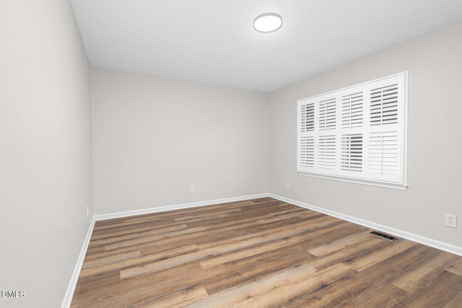 A small, empty room with light gray walls, white baseboards, wood-look vinyl flooring, a round ceiling light, and a window with white plantation shutters.