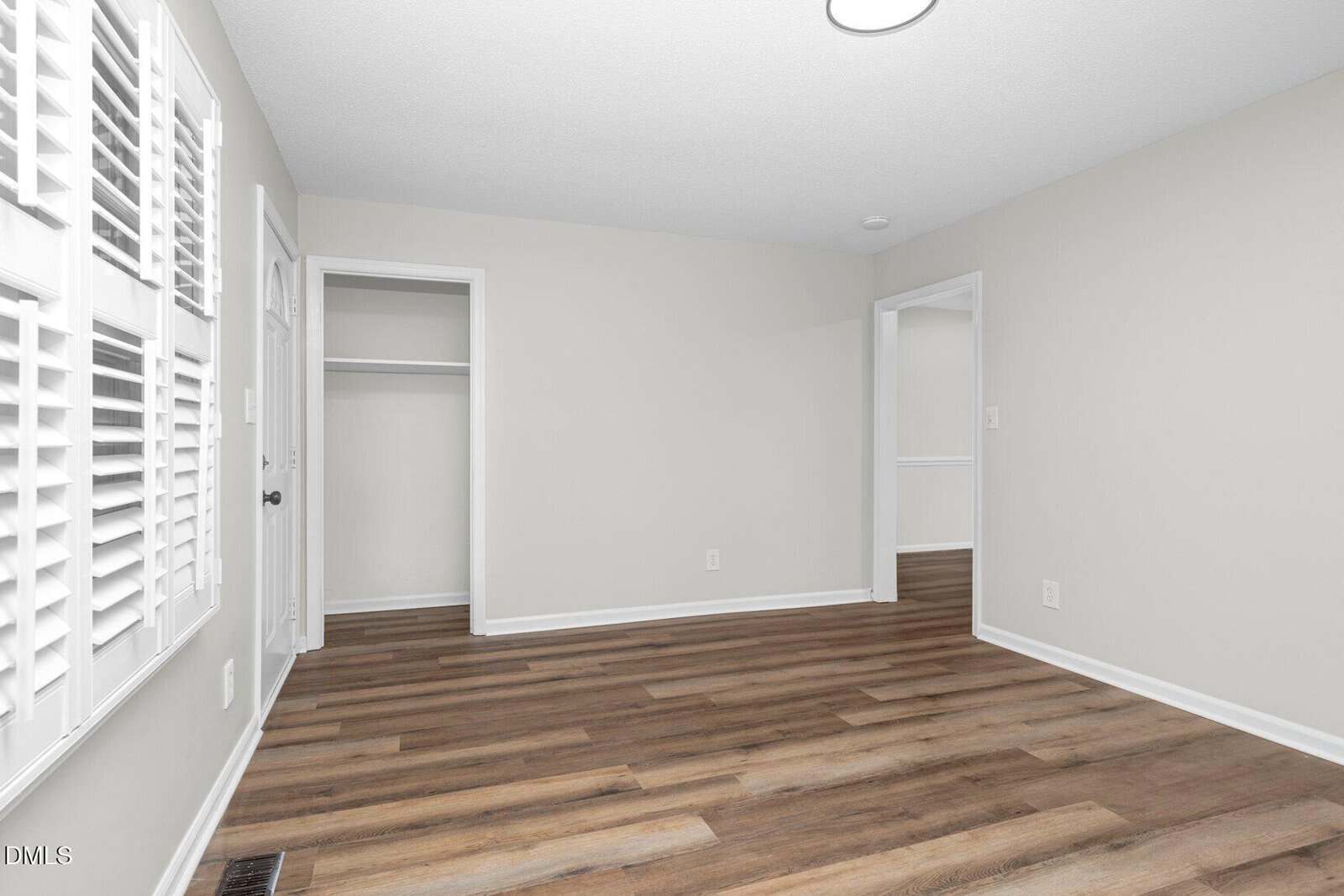 A clean, empty room with light gray walls, wood-look flooring, white trim, an open closet, two doorways, and multiple shuttered windows on the left letting in natural light.