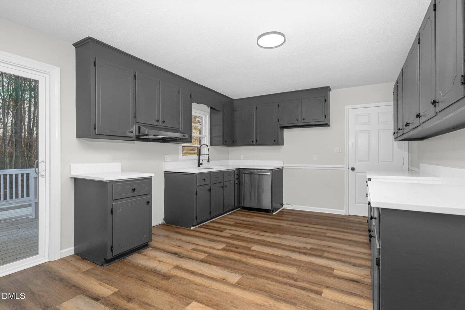 Modern kitchen with dark grey cabinets, white countertops, wood-look flooring, a large window over the sink, and a door leading to an outdoor deck. The room has neutral walls and recessed lighting.