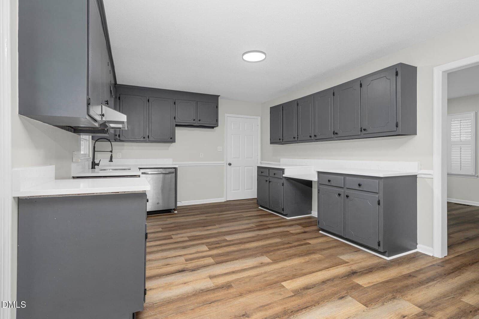 A modern kitchen with dark gray cabinets, white countertops, stainless steel appliances, wood-look flooring, and neutral-colored walls. There are multiple cabinets and drawers, and a door at the end of the room.