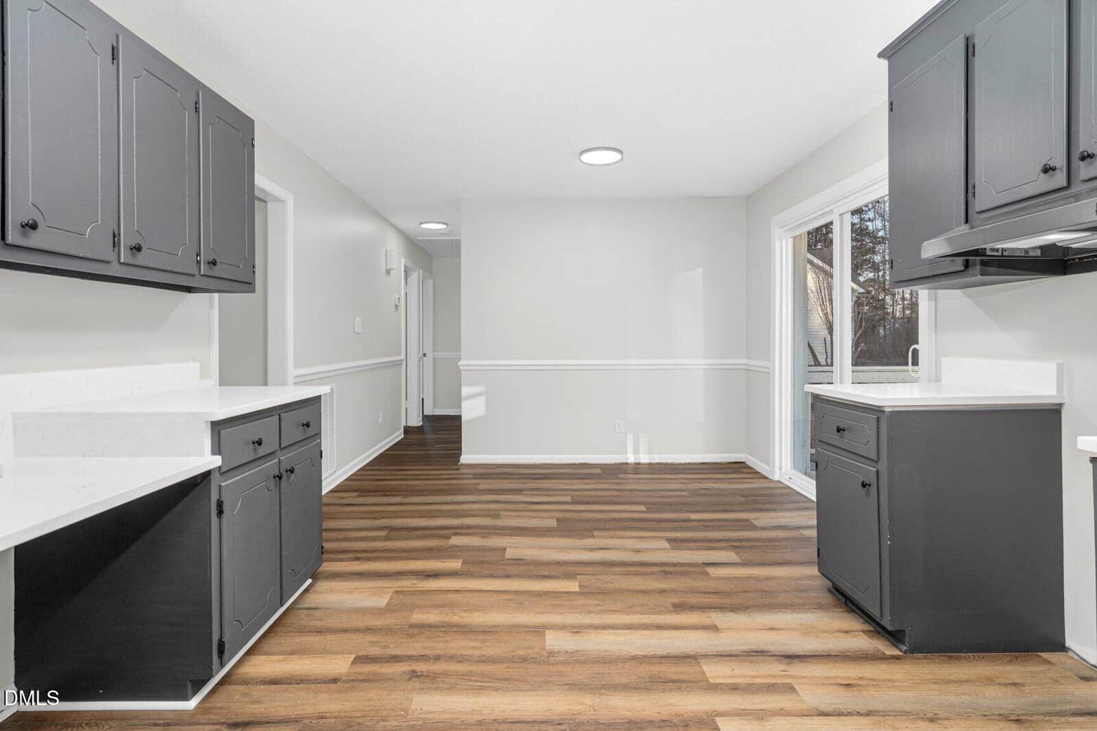 A kitchen with dark gray cabinets, white countertops, and wood-look flooring. The walls are light-colored, and there is a sliding glass door on the right that leads outside. The space is empty and well-lit.