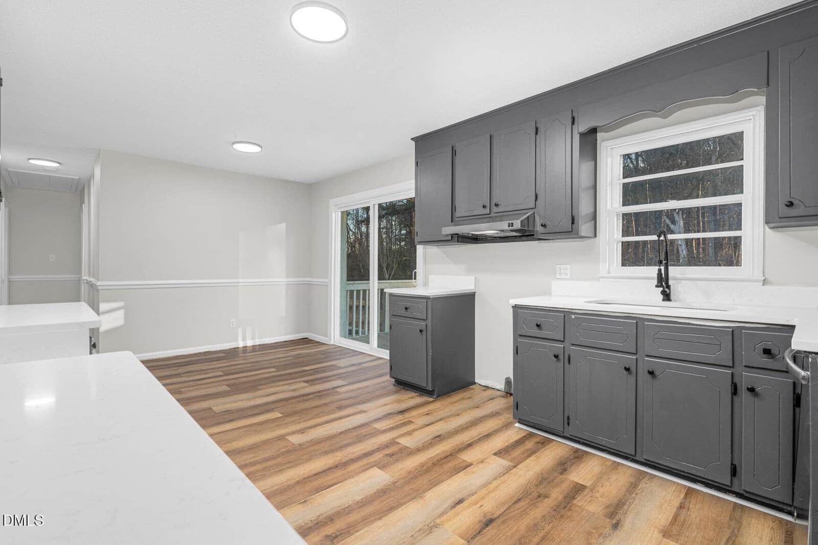 A kitchen with gray cabinets, white countertops, wood-look flooring, and a window above the sink. A sliding glass door leads outside, and the walls are painted light beige with white trim.