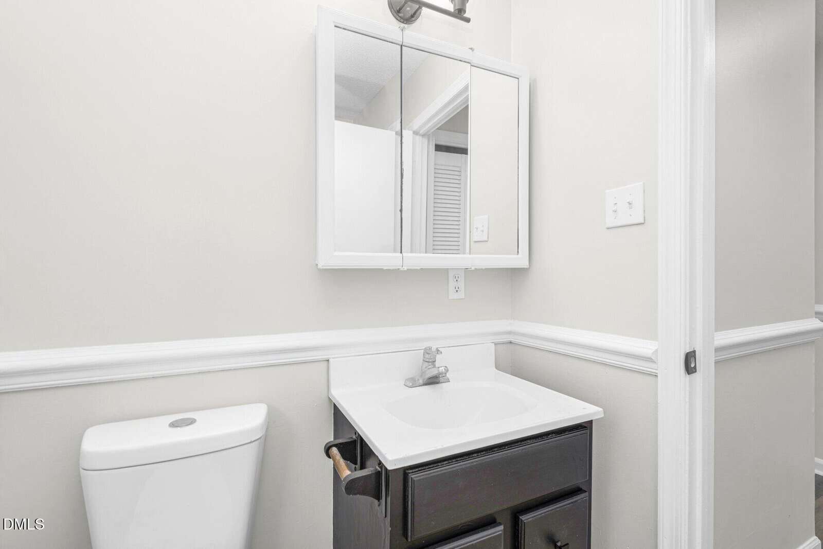 A small bathroom with a white toilet, dark vanity with a sink, mirrored medicine cabinet, and light gray walls with white trim.