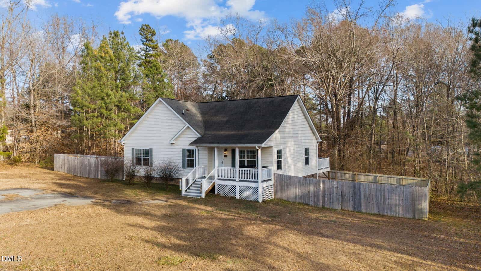 A single-story white house with a dark roof, front porch, and fenced backyard sits on a large, grassy lot. Leafless trees and pine trees surround the property under a partly cloudy sky.