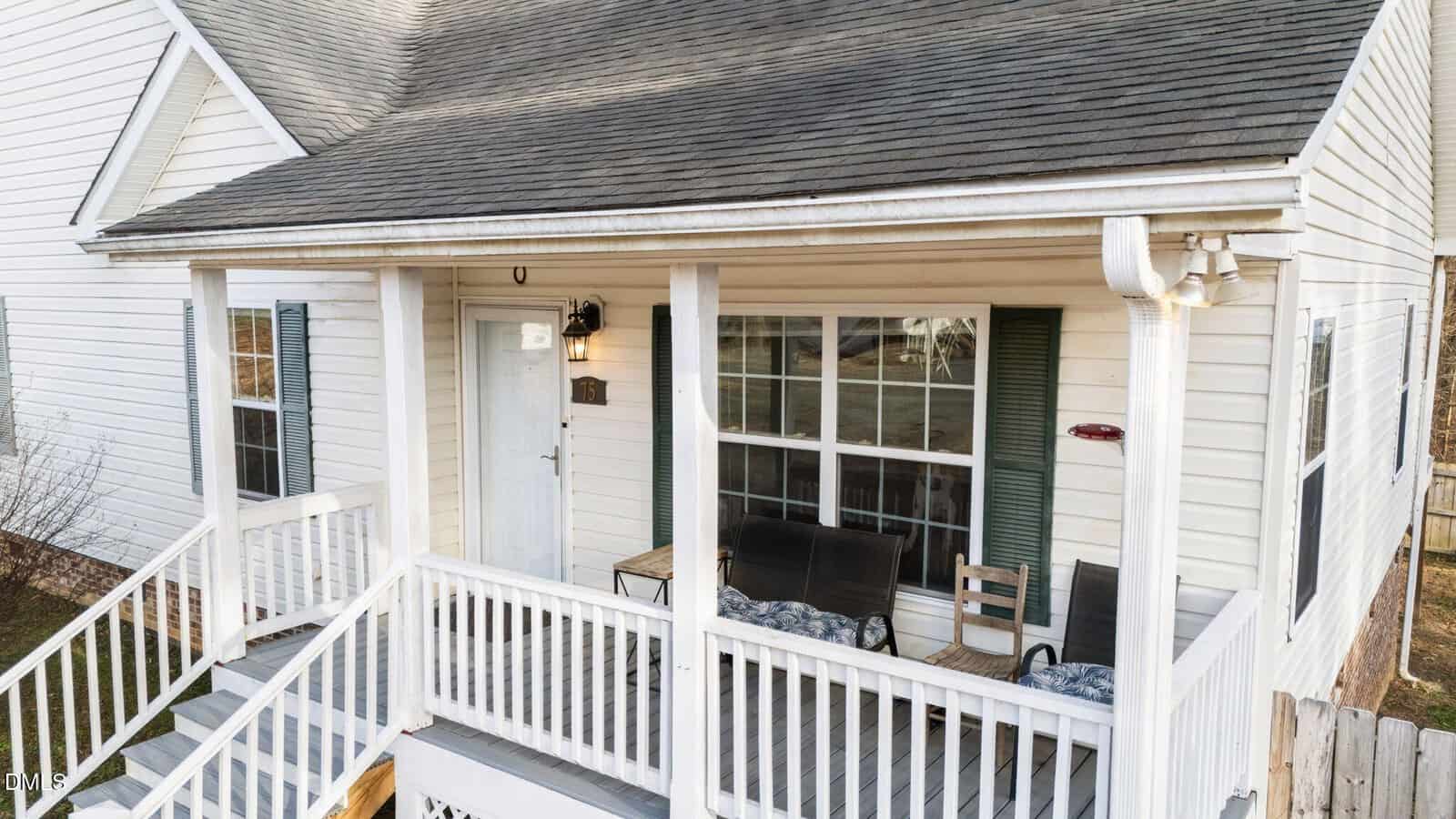 A white house with a covered front porch featuring a white railing, a light blue door, two green window shutters, outdoor seating, and steps leading down to the yard.