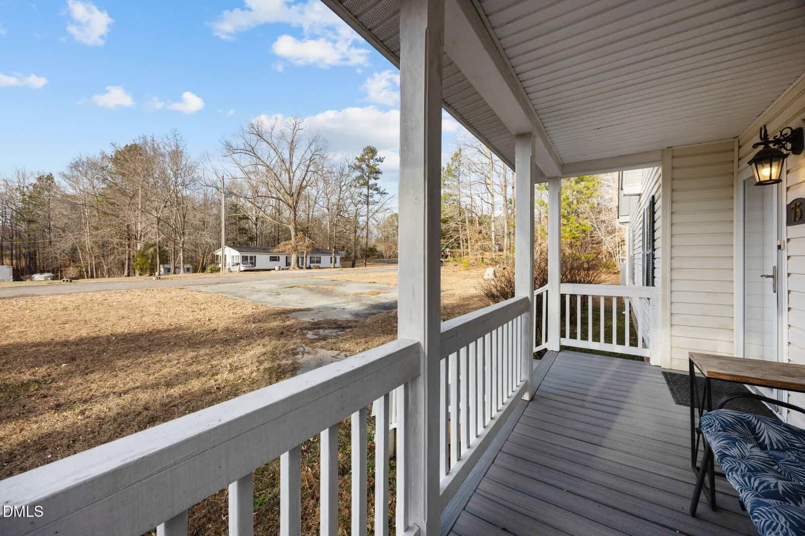 A view from a covered front porch with white railings, looking out over a dry lawn and driveway toward a distant house, with leafless trees and a partly cloudy sky in the background.