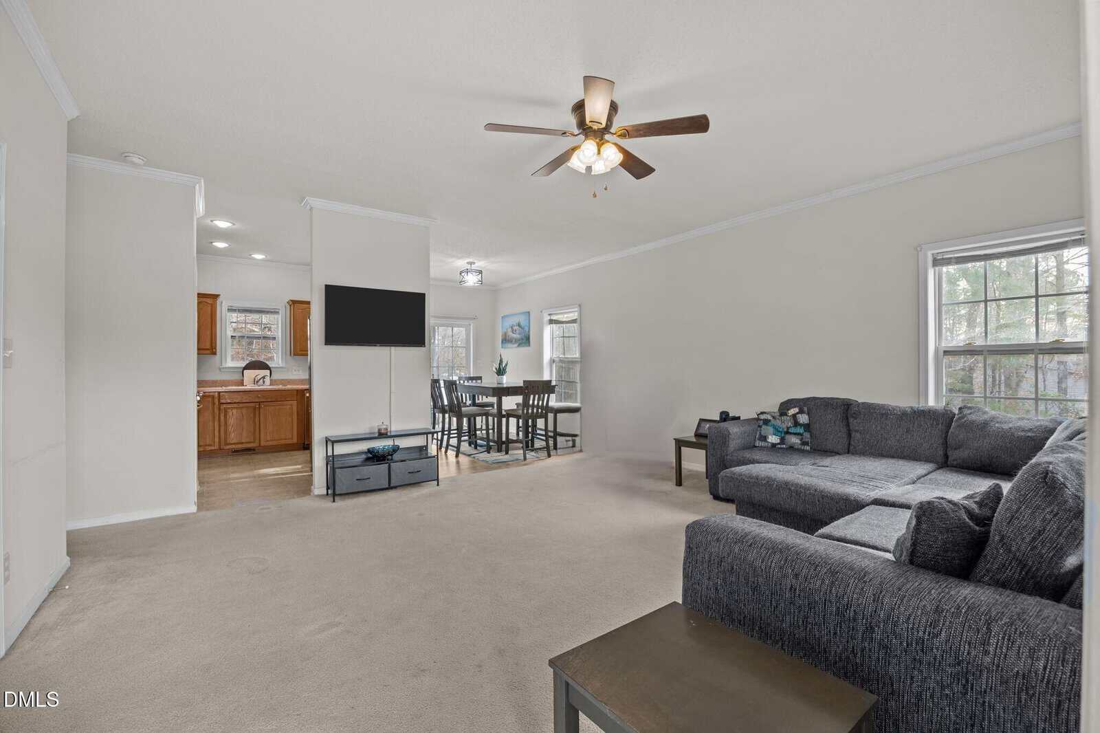 A spacious living room with beige carpet, a gray sectional sofa, a ceiling fan with lights, a flat-screen TV, and large windows. The space opens to a dining area and kitchen with wooden cabinets.