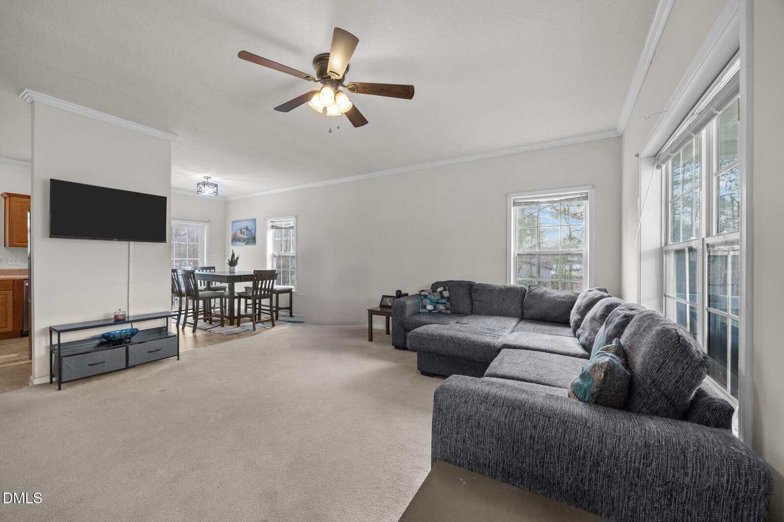 Spacious living room with a gray sectional sofa, large windows, beige carpet, ceiling fan, and a wall-mounted TV. Dining area with table and chairs is visible in the background.