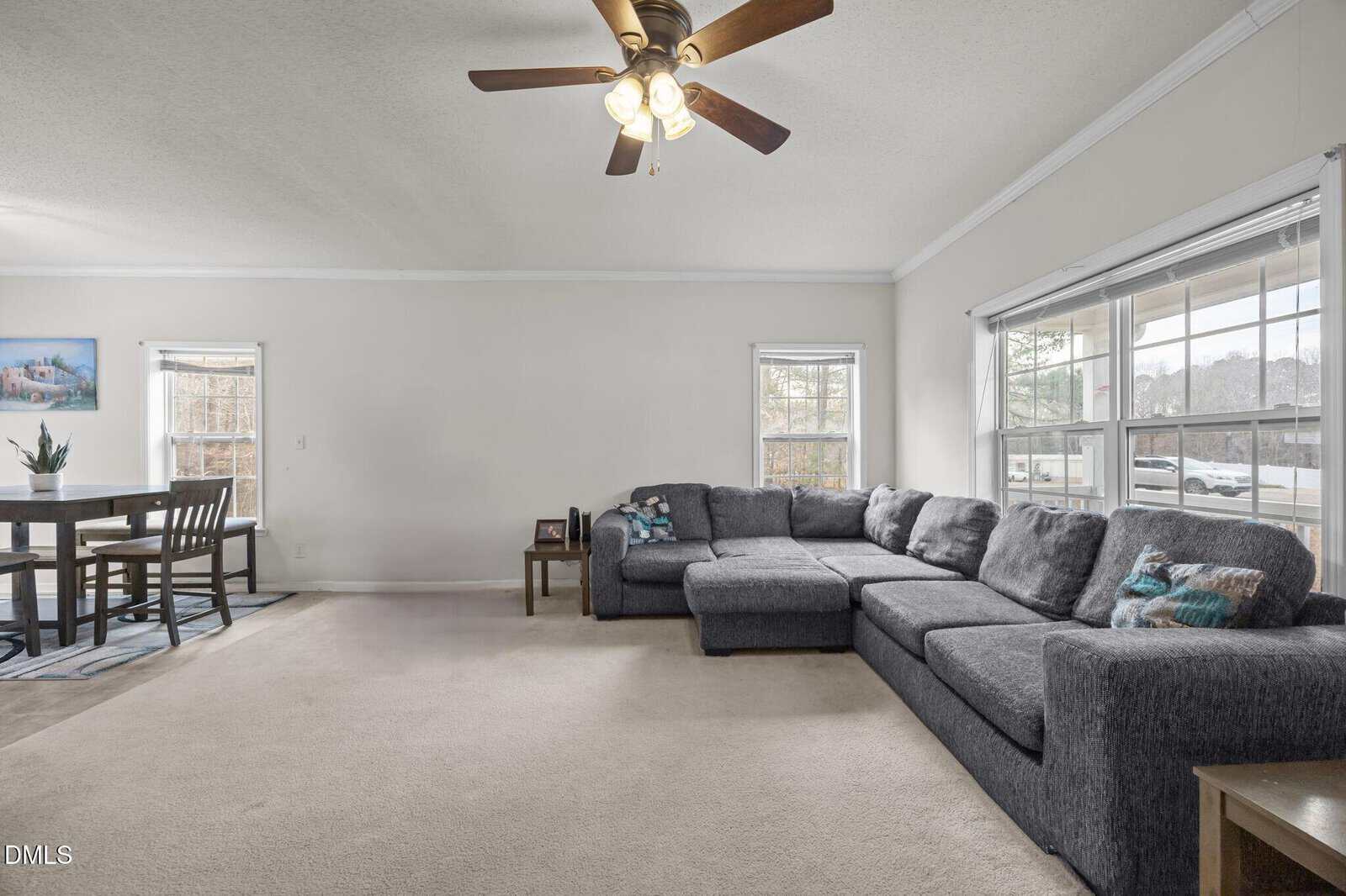 Spacious living room with a gray sectional sofa, side table, large windows, ceiling fan, and a dining area with a table and four chairs on a rug. Walls are light-colored and natural light fills the room.