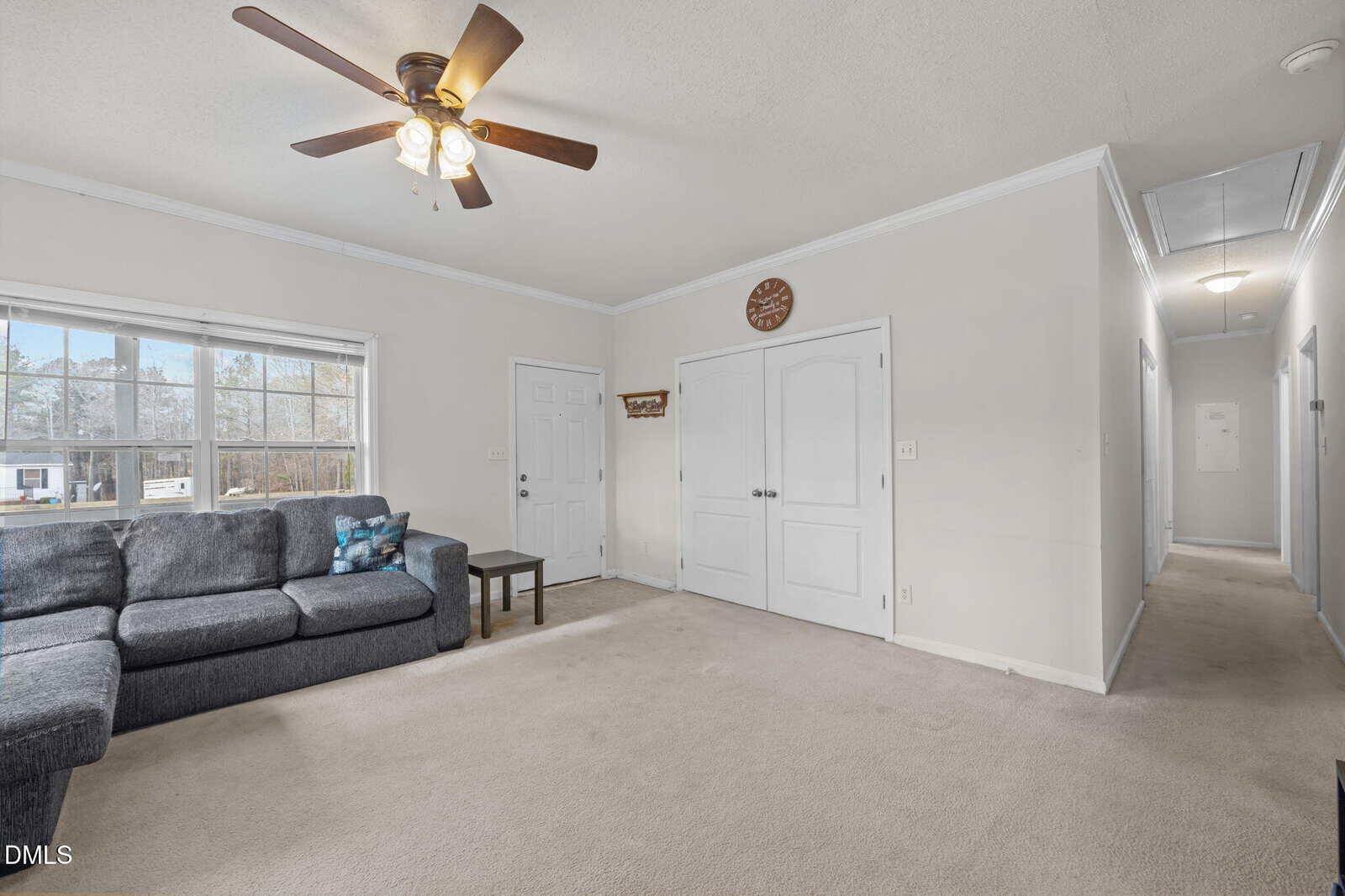 A living room with beige carpet, a gray sectional sofa, a small side table, a ceiling fan with lights, a double door closet, a window, and a hallway leading to other rooms.