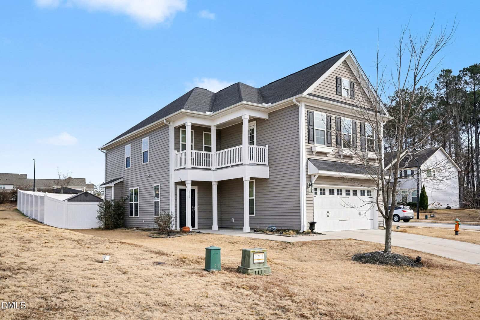 Two-story gray house with white trim, double garage, and small covered balcony. Dry grass lawn with a few small bushes and a leafless tree. White fence along the backyard. Blue sky and neighboring homes in the background.