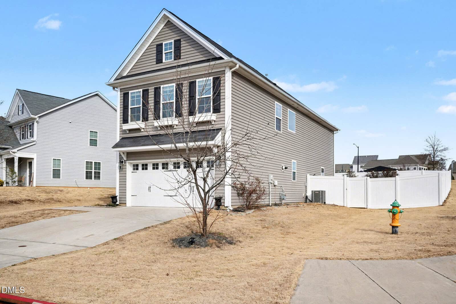 A two-story beige house with dark shutters and a white garage door, situated on a dry, brown lawn. A leafless tree is in the front yard, and a yellow fire hydrant is near the sidewalk. A white fence runs along the side.