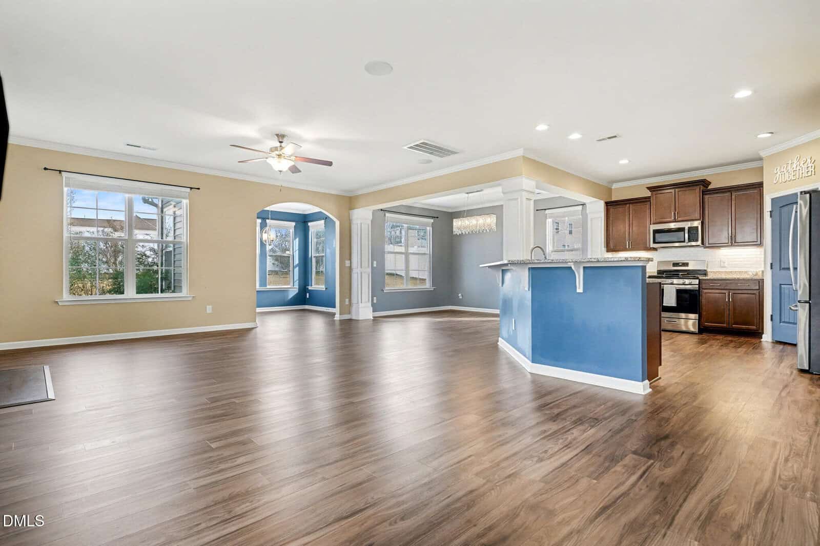 Open-plan living area with light wood flooring, a ceiling fan, large windows, blue accent walls, and an adjacent kitchen featuring dark wood cabinets, stainless steel appliances, and a breakfast bar.