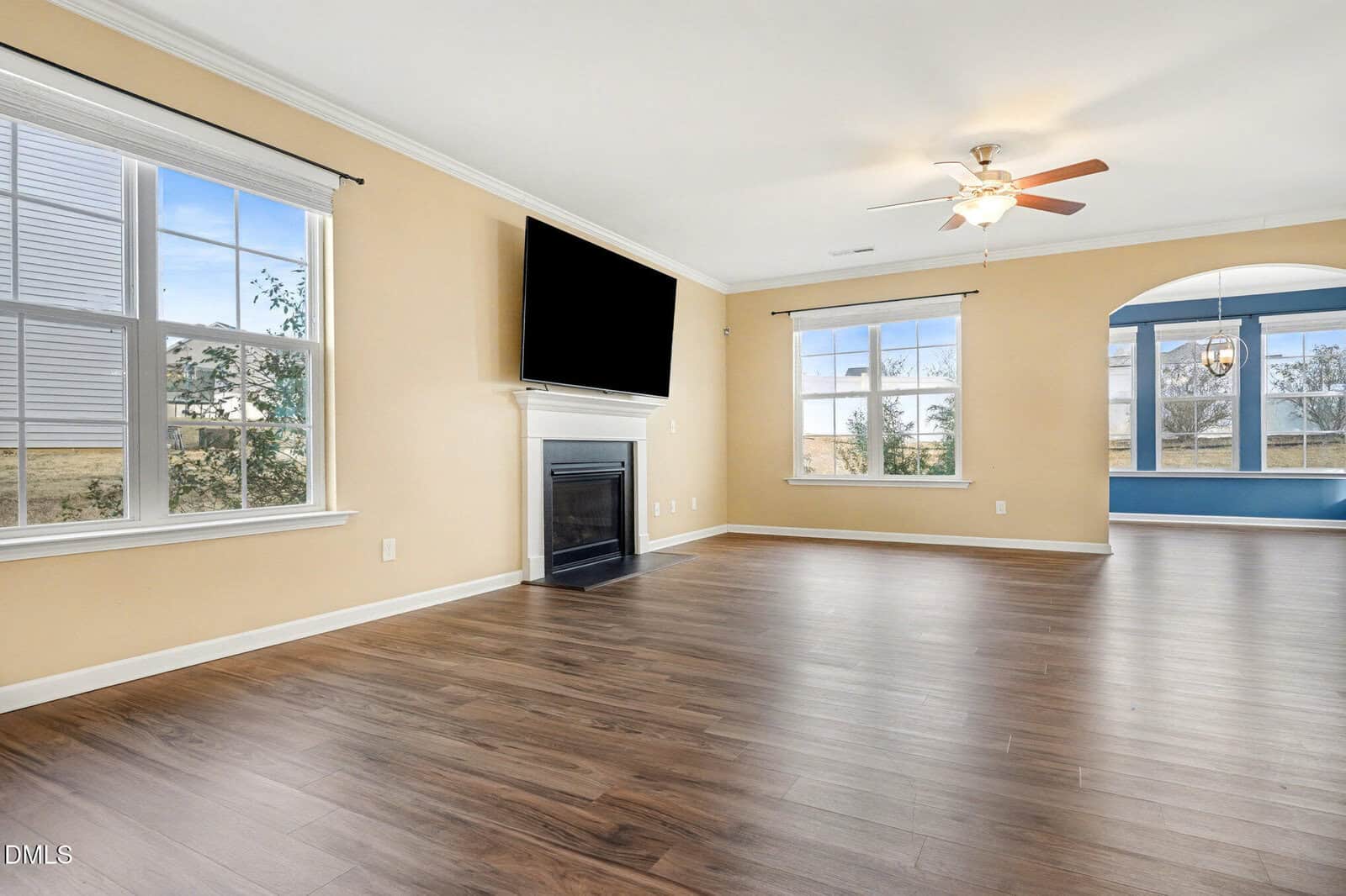 A spacious living room with light beige walls, large windows, wood flooring, a ceiling fan, a mounted TV above a fireplace, and an adjacent dining area with blue accent walls.