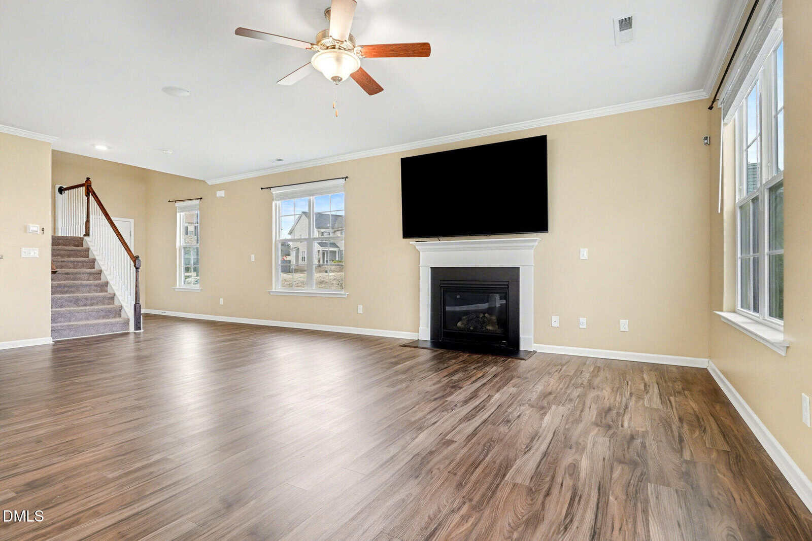 Spacious living room with wood flooring, beige walls, large windows, a fireplace with a TV above it, and a ceiling fan. A staircase is visible on the left side of the room.