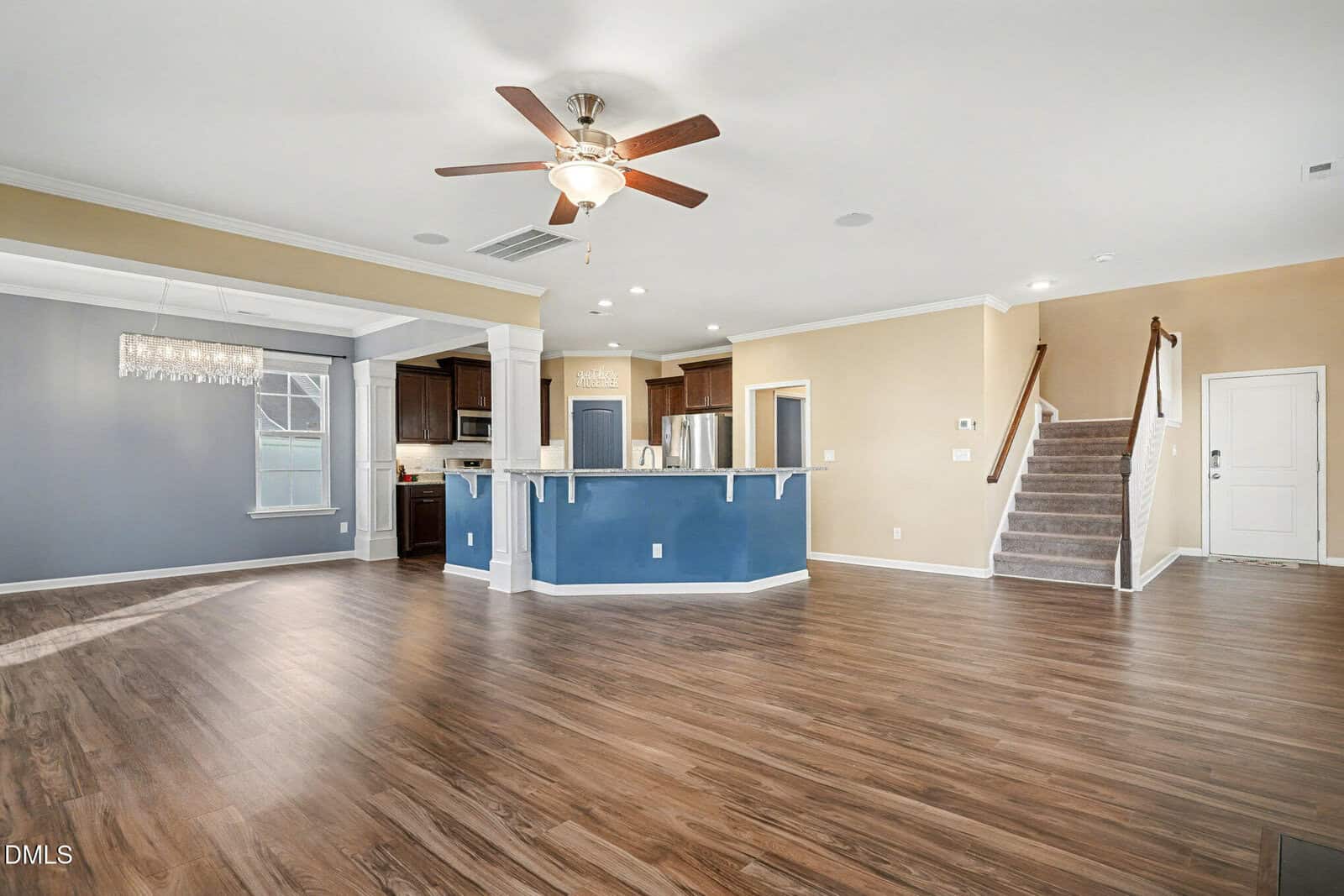 Spacious open-concept living area with wood-look flooring, a ceiling fan, a blue kitchen island, stainless steel appliances, a dining area with a chandelier, and stairs leading to the upper level.