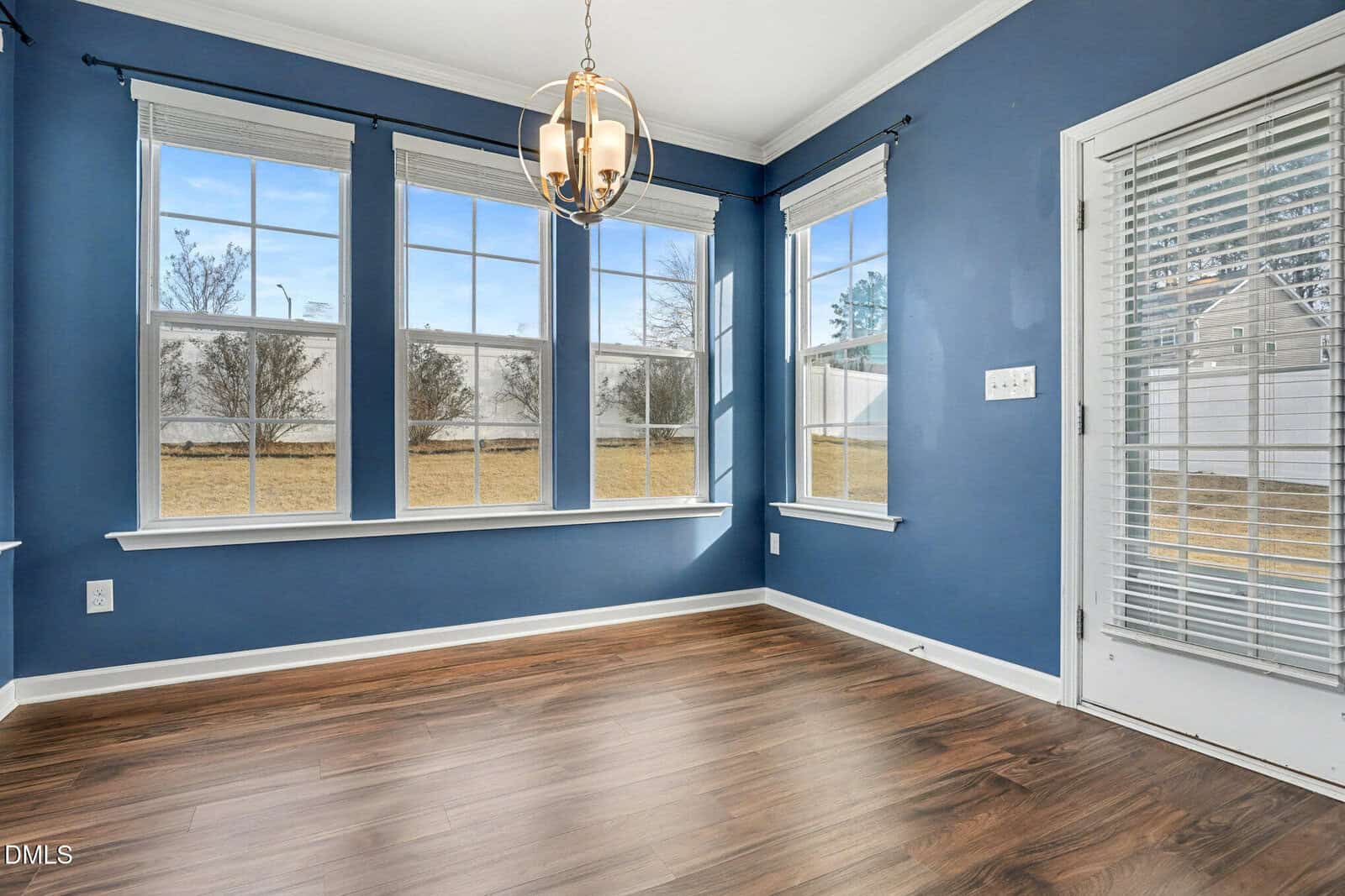 A room with blue walls, three large windows with white trim, wood flooring, and a glass door with blinds. A modern chandelier hangs from the ceiling. The windows overlook a grassy outdoor area.