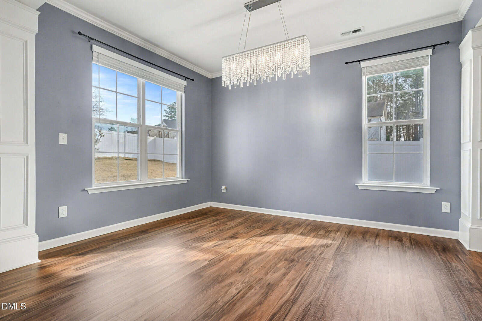 An empty room with light purple-gray walls, two large windows, wood flooring, white trim, and a modern rectangular crystal chandelier hanging from the ceiling.