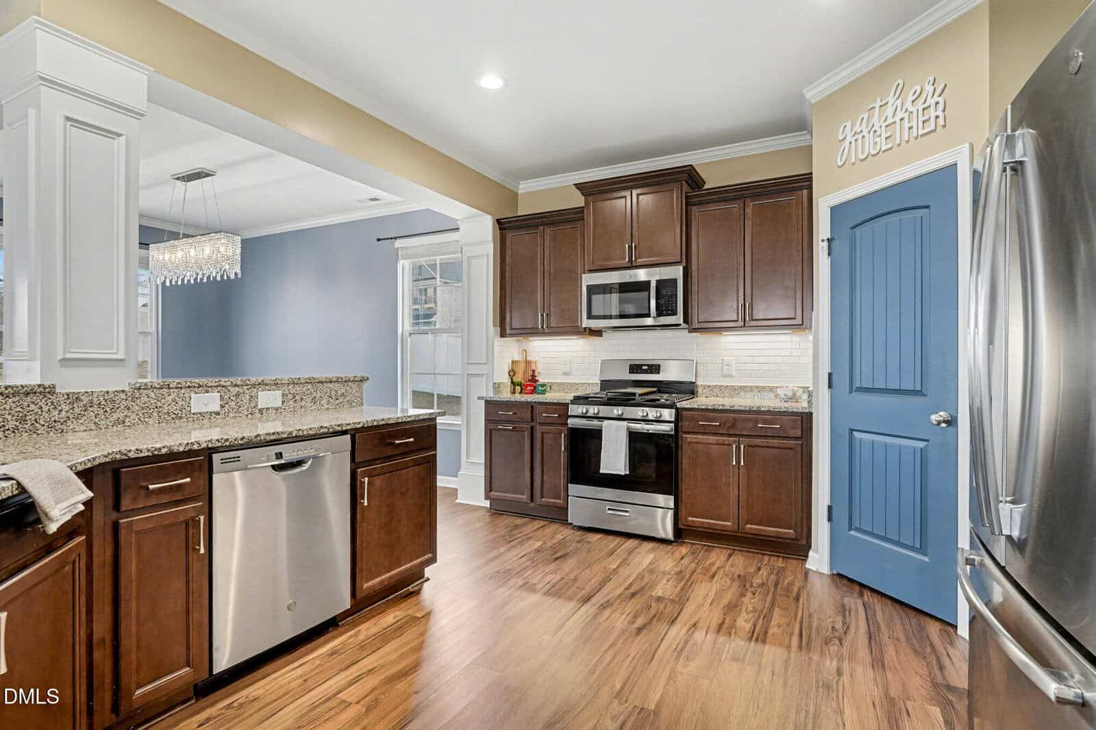 Modern kitchen with stainless steel appliances, brown cabinets, granite countertops, wood flooring, and a blue pantry door. Dining area with chandelier visible in the background. Bright, clean, and spacious design.