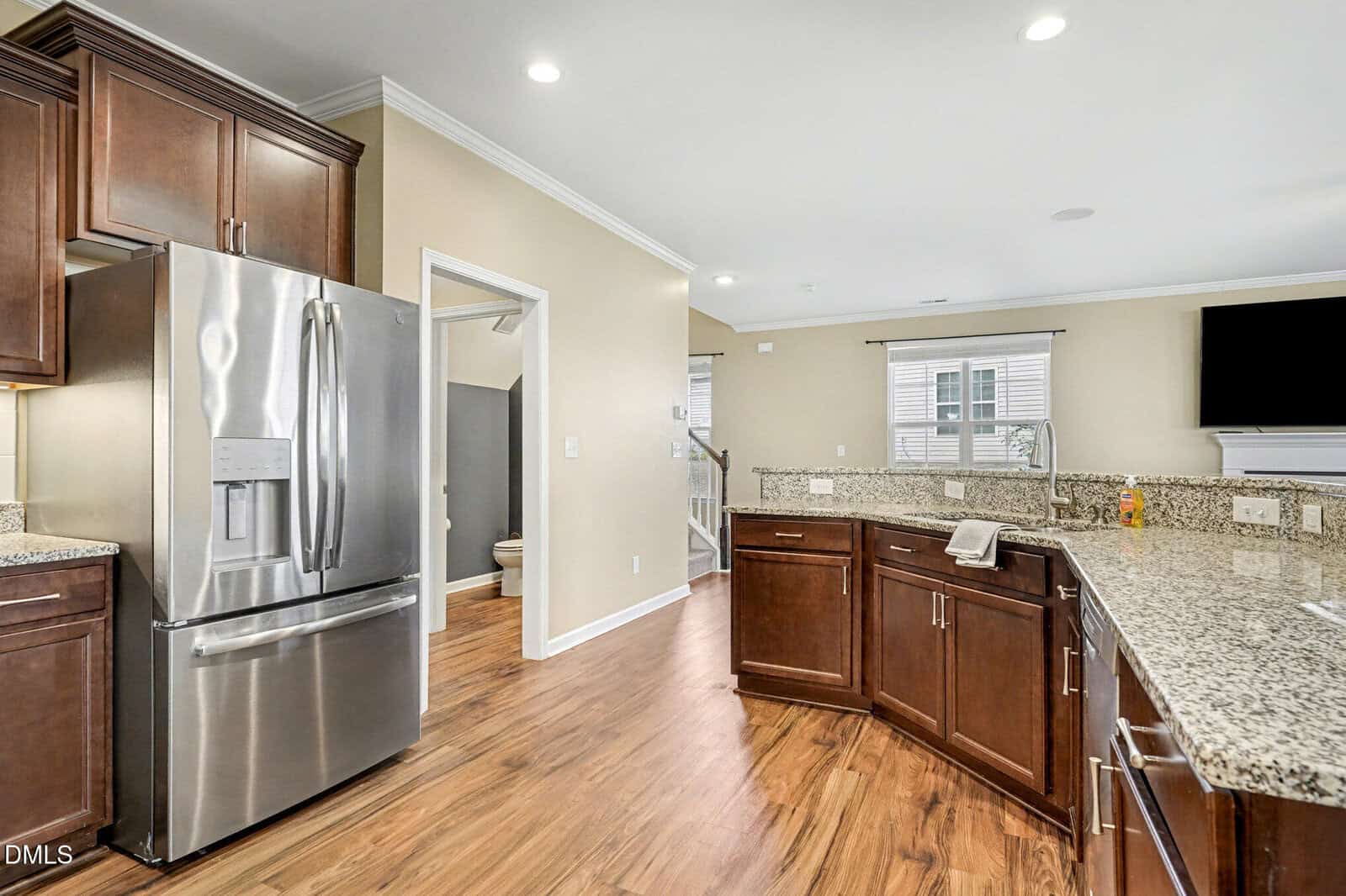 Modern kitchen with stainless steel refrigerator, brown cabinets, granite countertops, wood flooring, and a view of an adjacent bathroom and living area in the background.