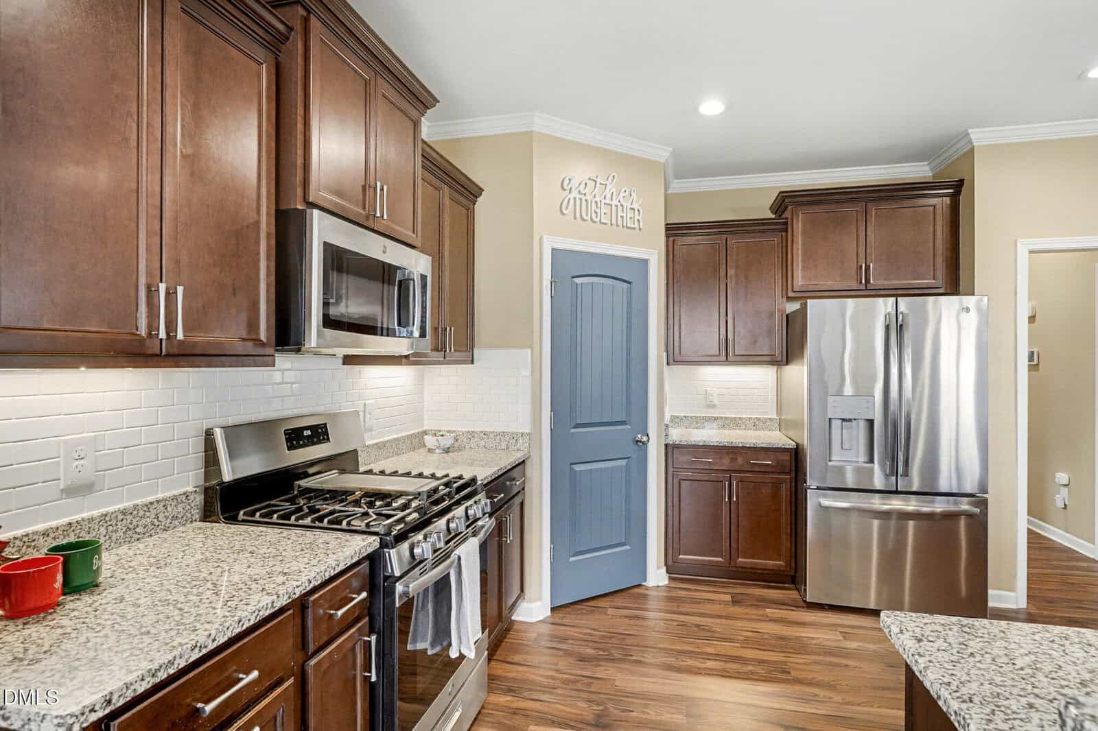 Modern kitchen with stainless steel appliances, granite countertops, brown cabinets, white subway tile backsplash, and a blue pantry door. A “Gather Together” sign is above the door.