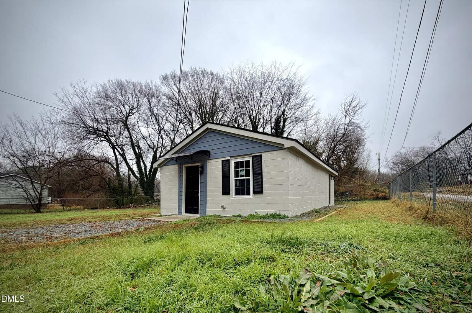 A small, single-story building with gray siding and white trim sits on a grassy lot. There are leafless trees in the background and a chain-link fence on the right side. The sky is overcast.