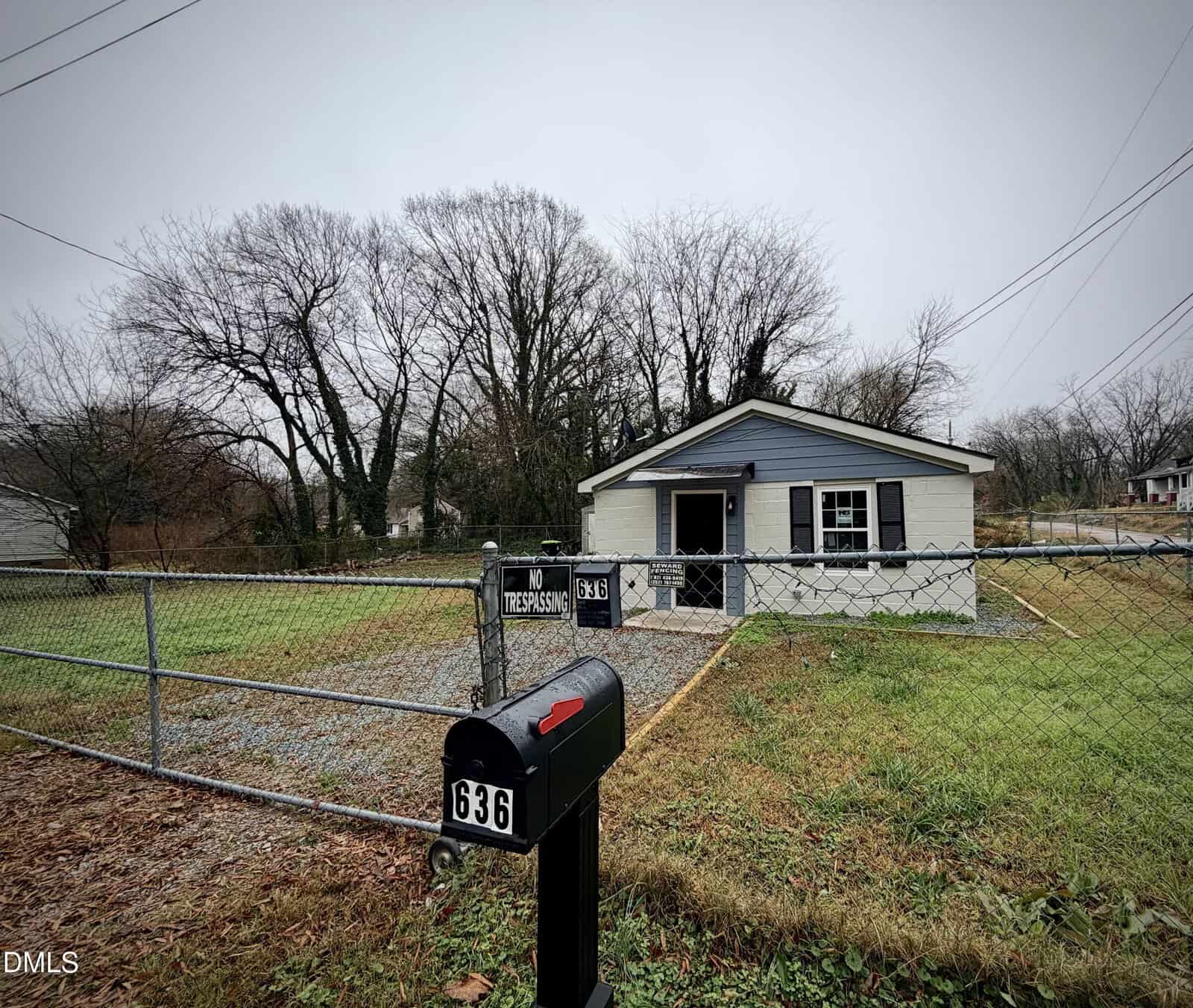 A small, single-story house with gray siding and white trim is behind a chain-link fence. A "No Trespassing" sign and the number 636 are visible on the fence and mailbox. The yard has patchy grass and leafless trees.