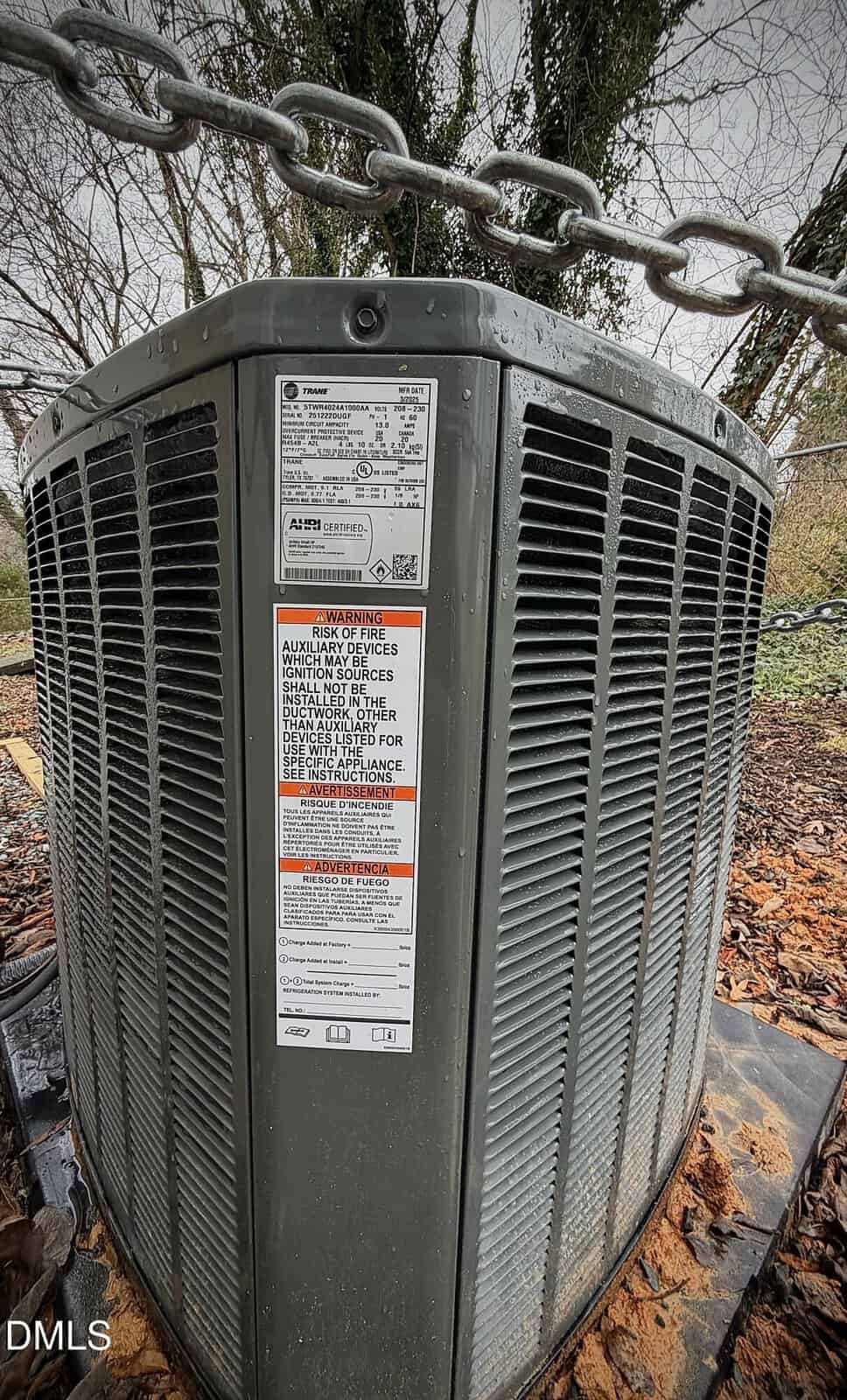 Close-up view of a large outdoor air conditioning unit with warning and information labels, surrounded by wet leaves and a chain overhead. Trees and overcast sky are visible in the background.