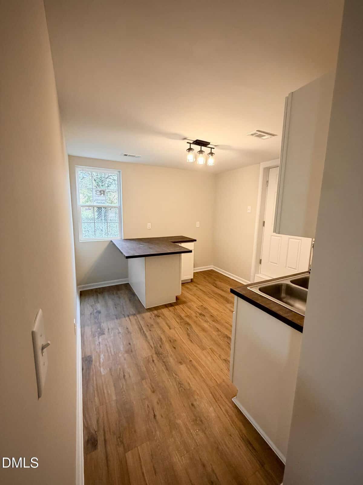 A small kitchen with wood-style flooring, white walls, a window, brown countertops, and a ceiling light fixture. There is a counter area with an L-shaped layout and an under-mount sink on the right side.