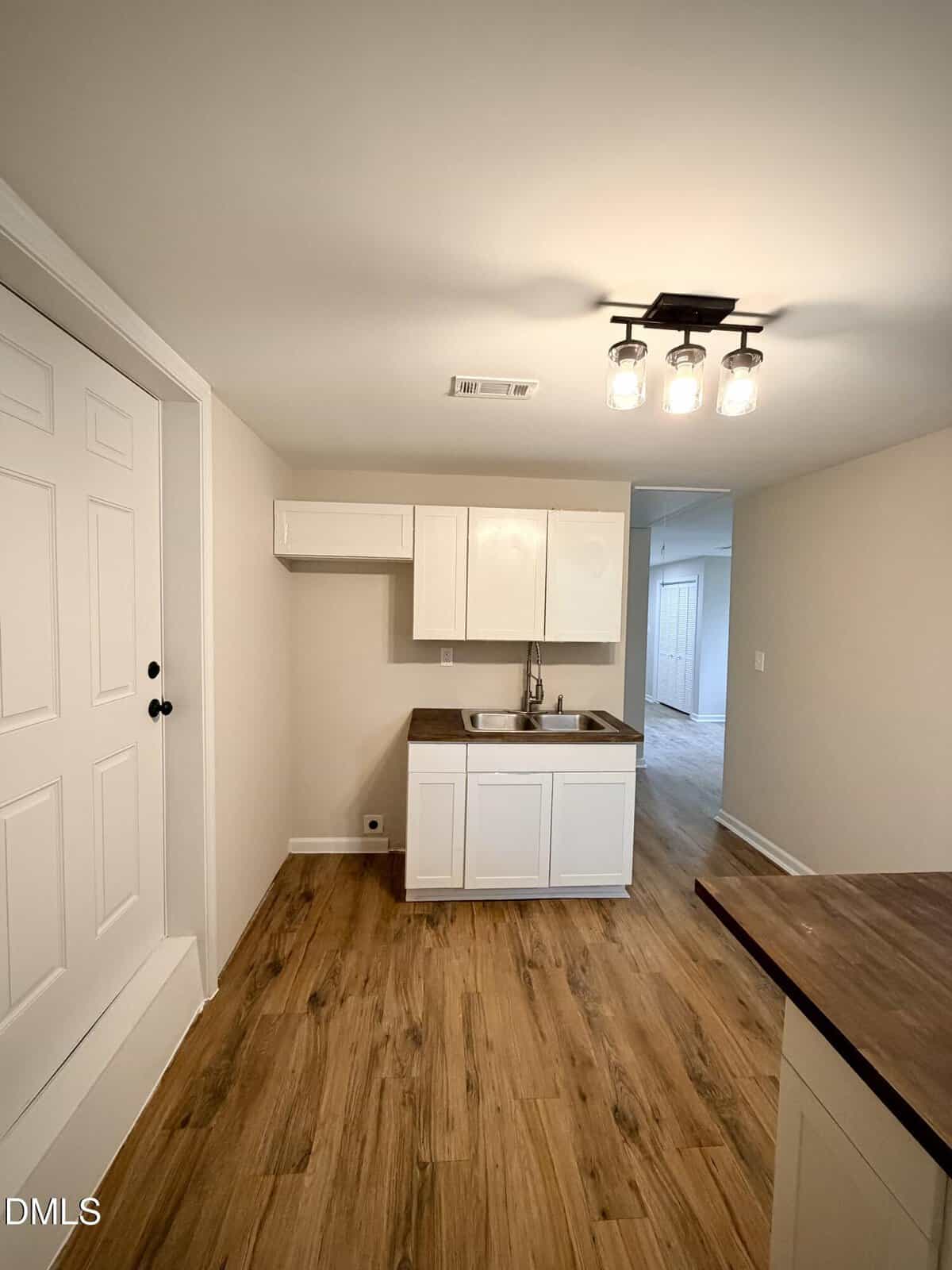 A small kitchen area with white cabinets, a stainless steel sink, brown countertops, wood-look flooring, and a ceiling light fixture. A white door is on the left, and a hallway is visible in the background.