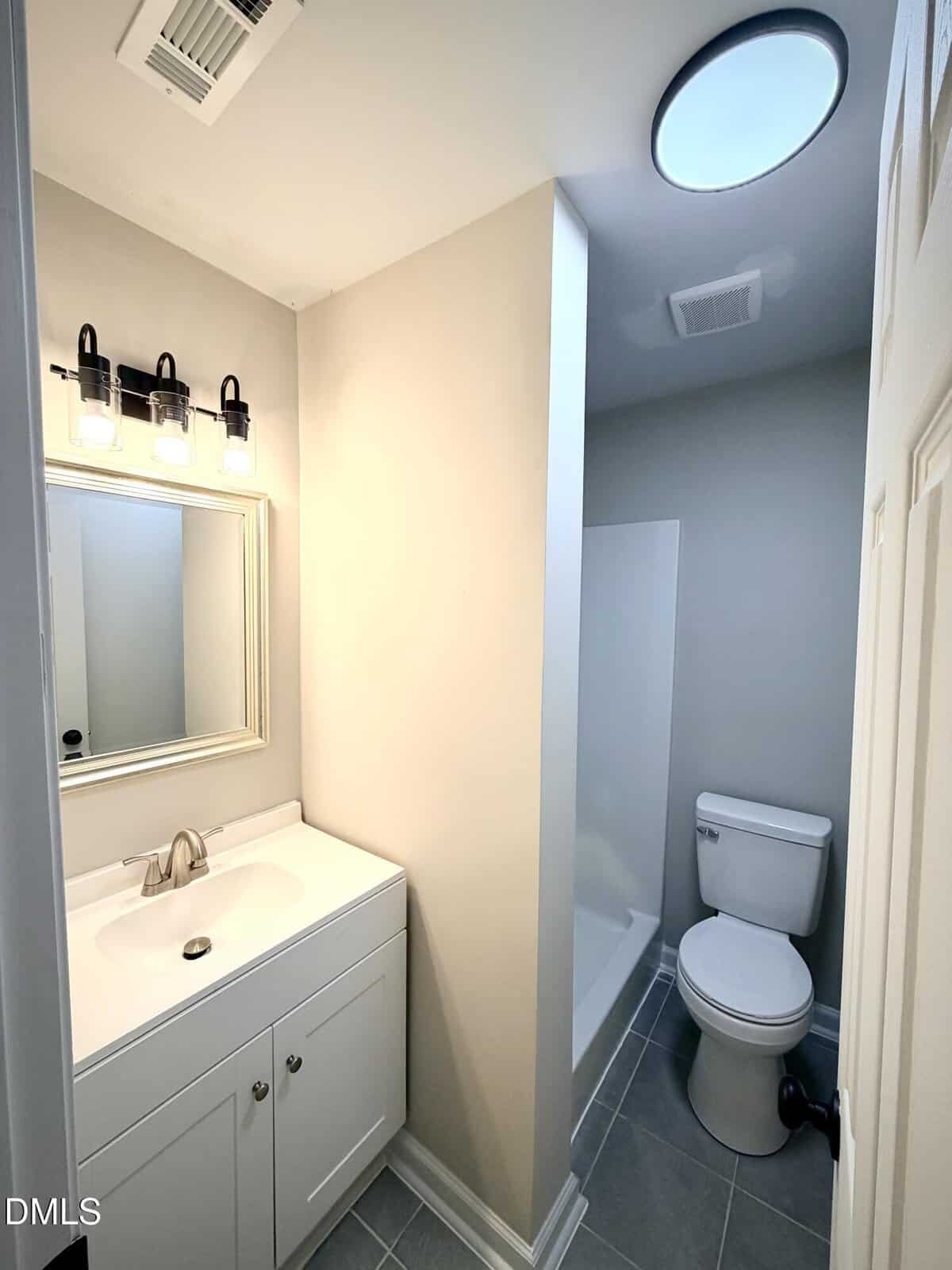 A small bathroom with a white vanity, sink, and mirror on the left, gray tile floor, a white toilet, and a shower area separated by a partial wall. Ceiling lights and neutral wall colors are present.