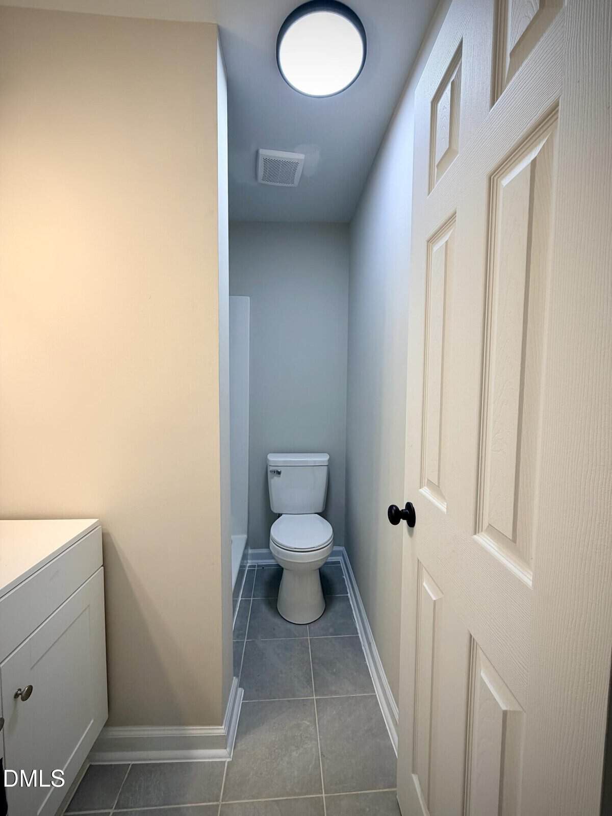 A small bathroom with gray tile floor, white walls, a white toilet at the end of a narrow alcove, and part of a white vanity and door visible. Ceiling light fixture above.