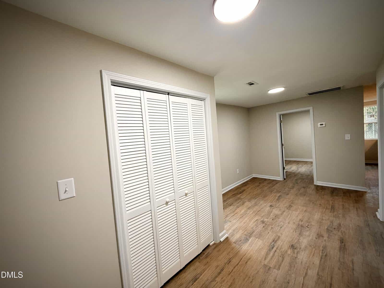 A room with light beige walls, wood-style flooring, a white double-door closet, and a ceiling light. An open doorway leads to another room with similar flooring and wall color.