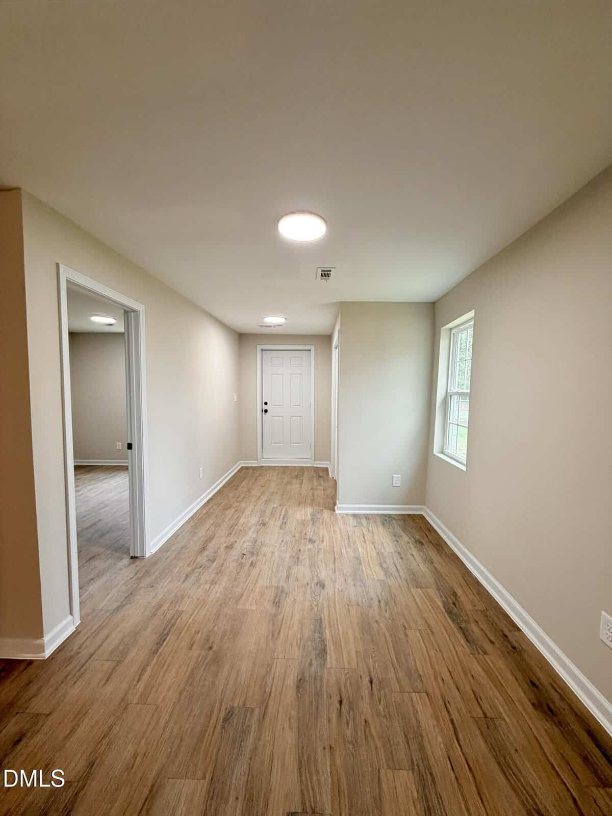A narrow hallway with wood-look flooring, beige walls, white trim, two windows on the right, open doorways on the left, and a white entry door at the far end. Ceiling lights are installed overhead.